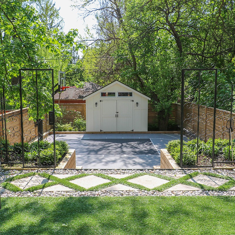 A small white shed is surrounded by trees and a well-maintained garden, with stone pathways leading to a black metal gate.