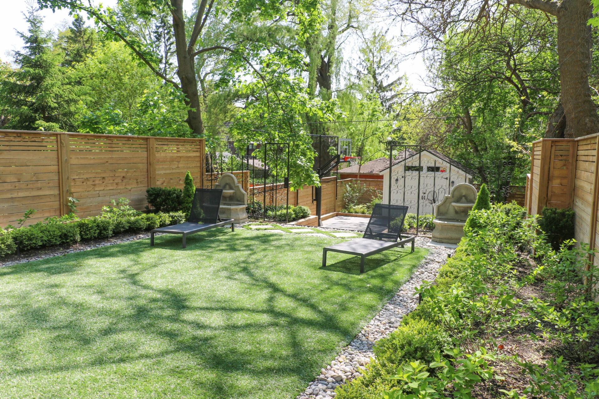 A serene backyard with lush greenery, lawn chairs on artificial grass, wooden fencing, and a small shed in a cozy setting.