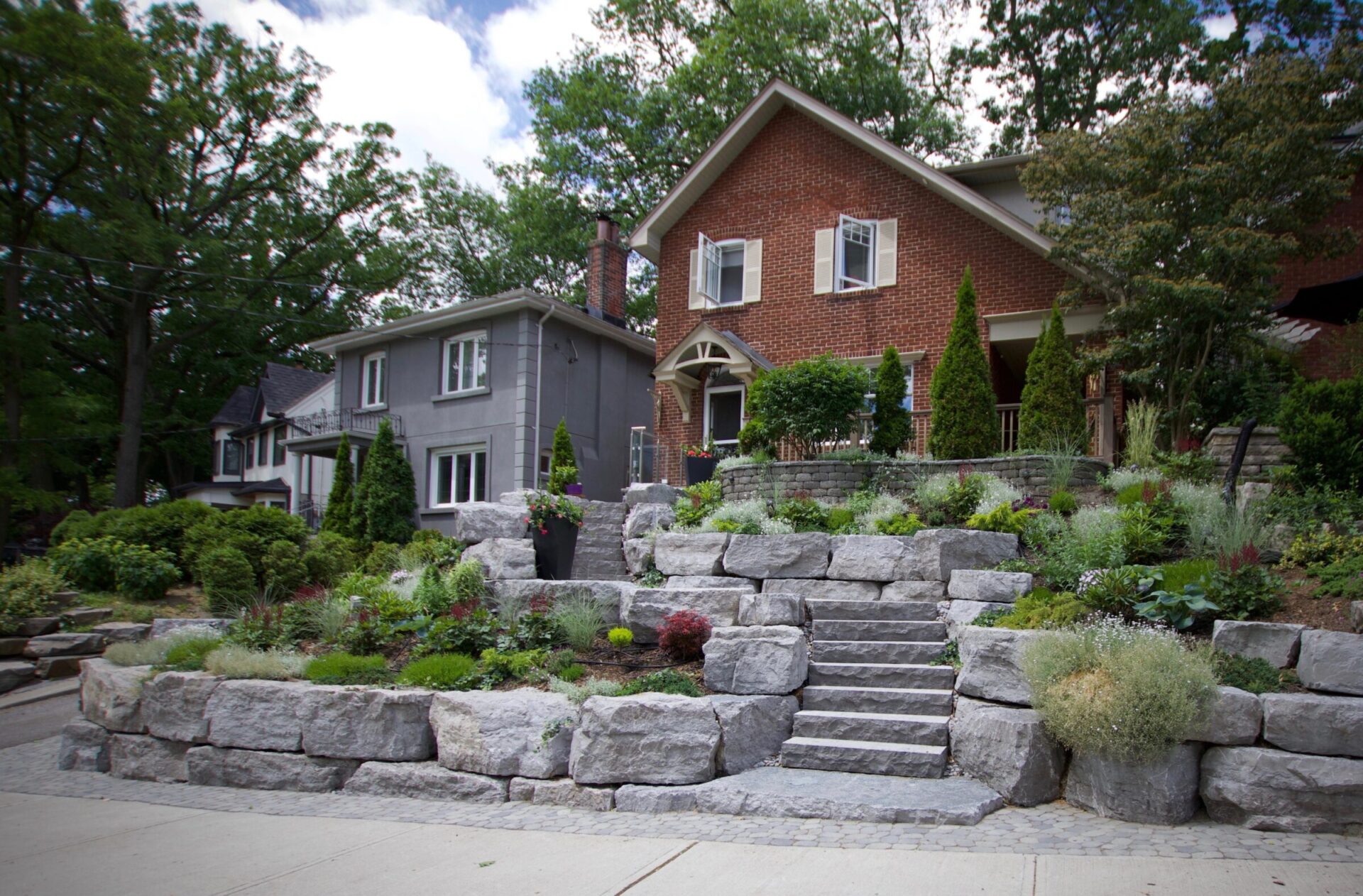 Two suburban houses with lush gardens, stone retaining walls, and stairs. Trees in the background enhance the peaceful neighborhood setting.