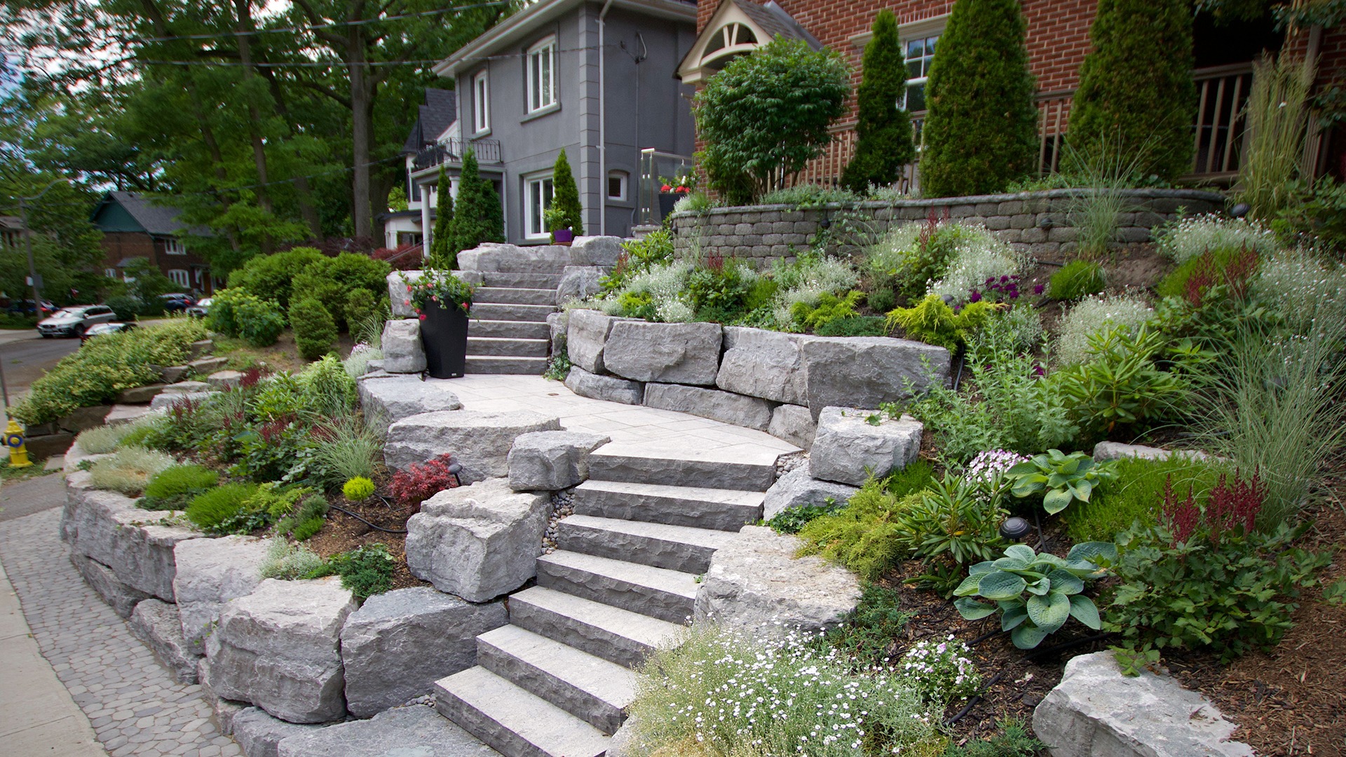 A landscaped garden features stone steps and lush greenery, with residential houses in the background, creating a picturesque and tranquil suburban scene.