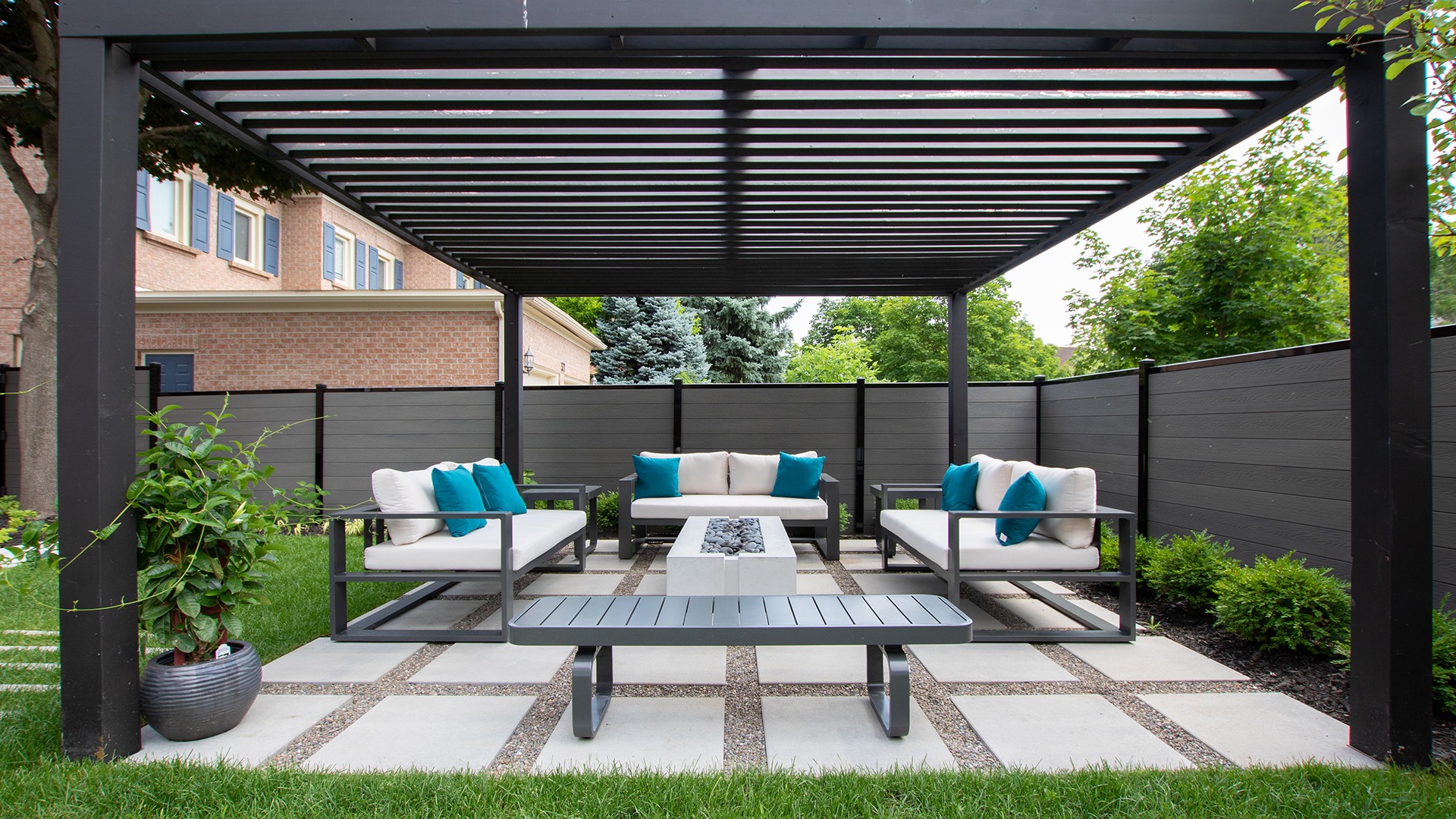 Outdoor patio with modern furniture, teal pillows, under pergola. Surrounded by greenery and brick wall, creating a cozy and inviting atmosphere.