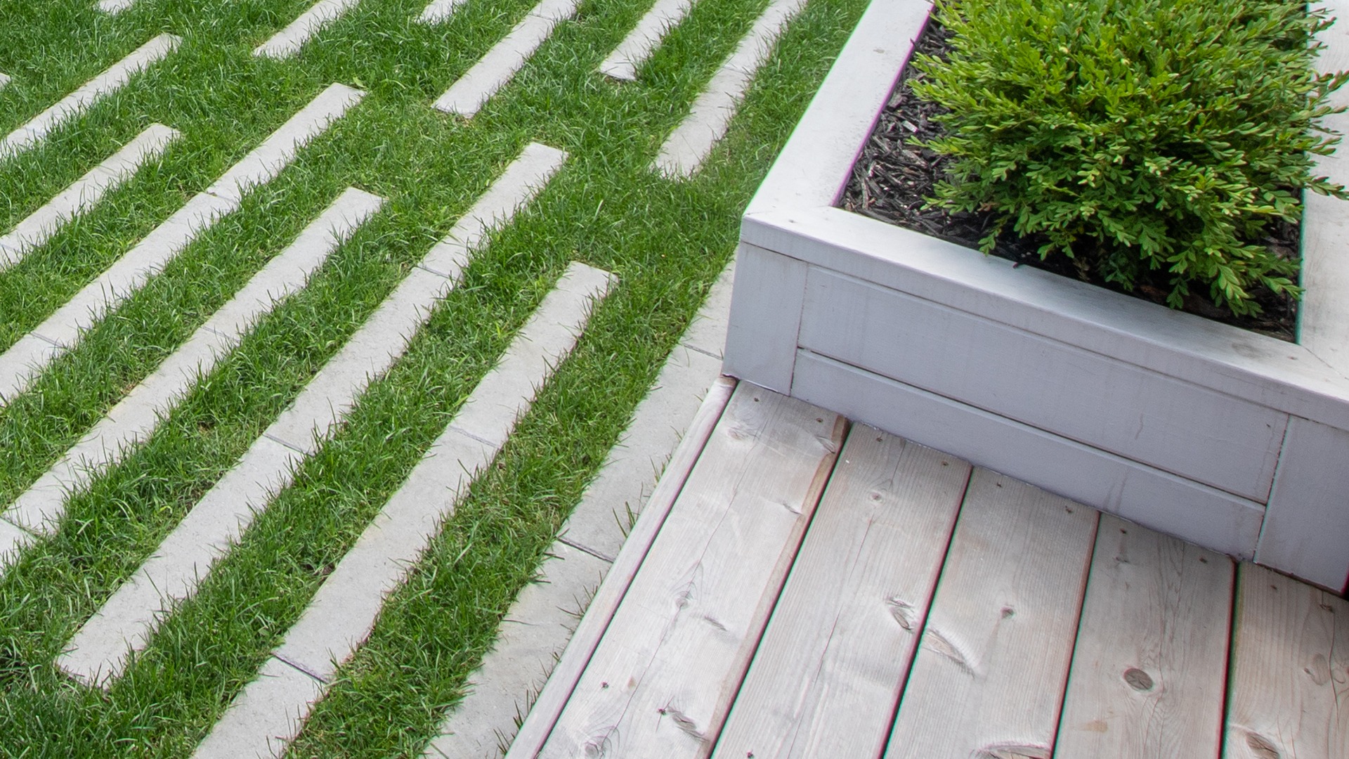 A modern garden features a wooden deck, linear stone path, and rectangular planter box with lush green shrub, creating a minimalist outdoor space.
