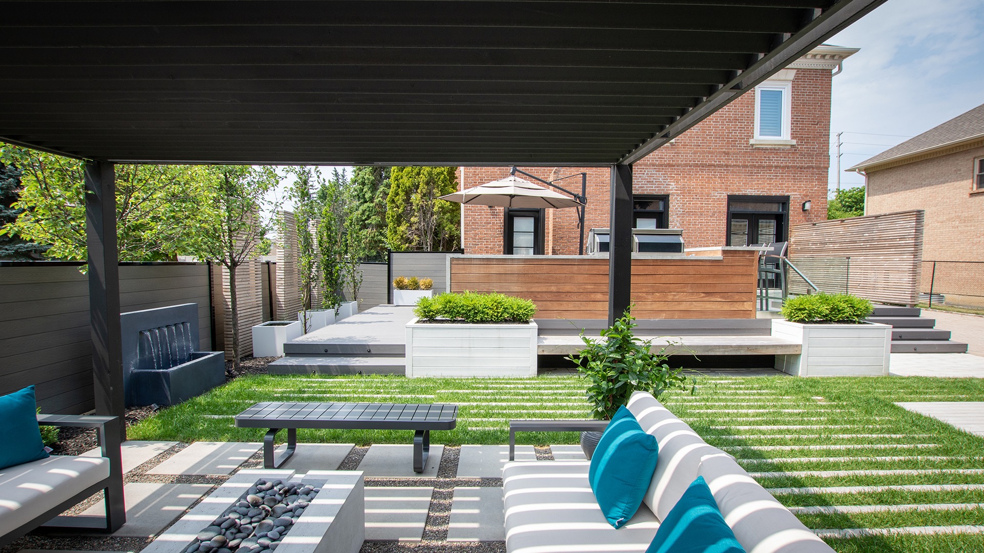 Modern backyard with pergola, seating, and greenery. Features a brick house, wooden deck, umbrella, and stone pathway, creating a relaxed outdoor space.