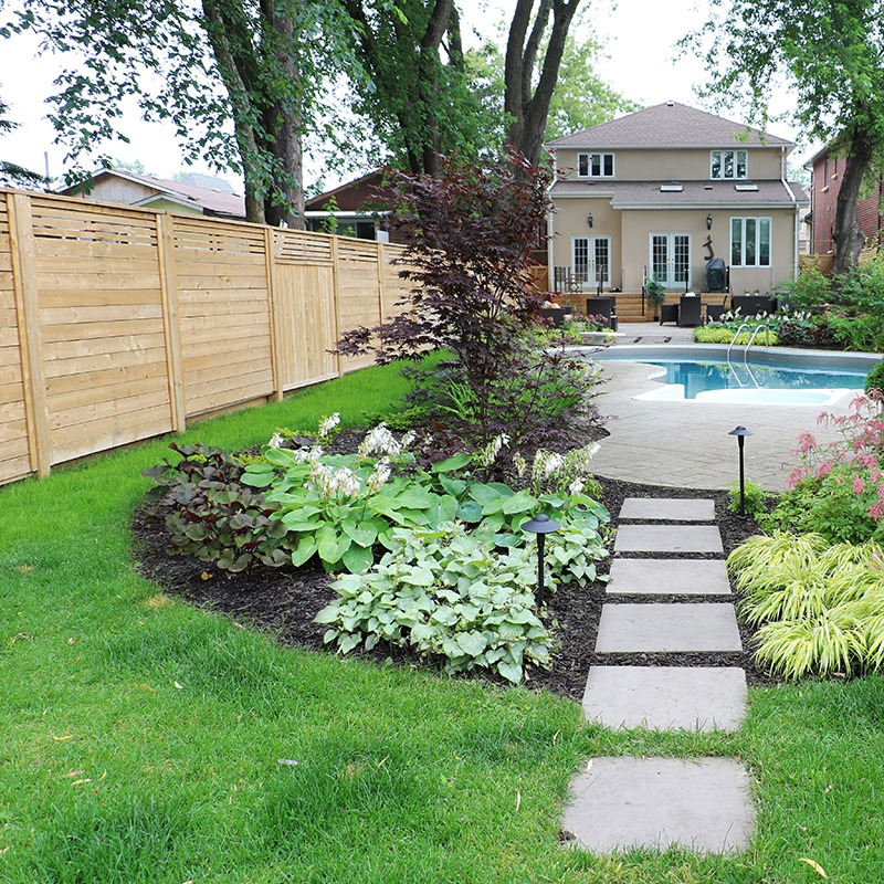 A well-maintained backyard features a pool, lush greenery, patio area, wooden fencing, and a stepping stone path leading up to a beige house.