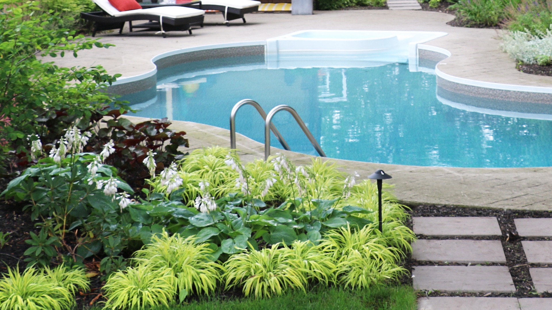 A curved swimming pool surrounded by lush green plants and flowers, with lounge chairs in the background on a stone patio.