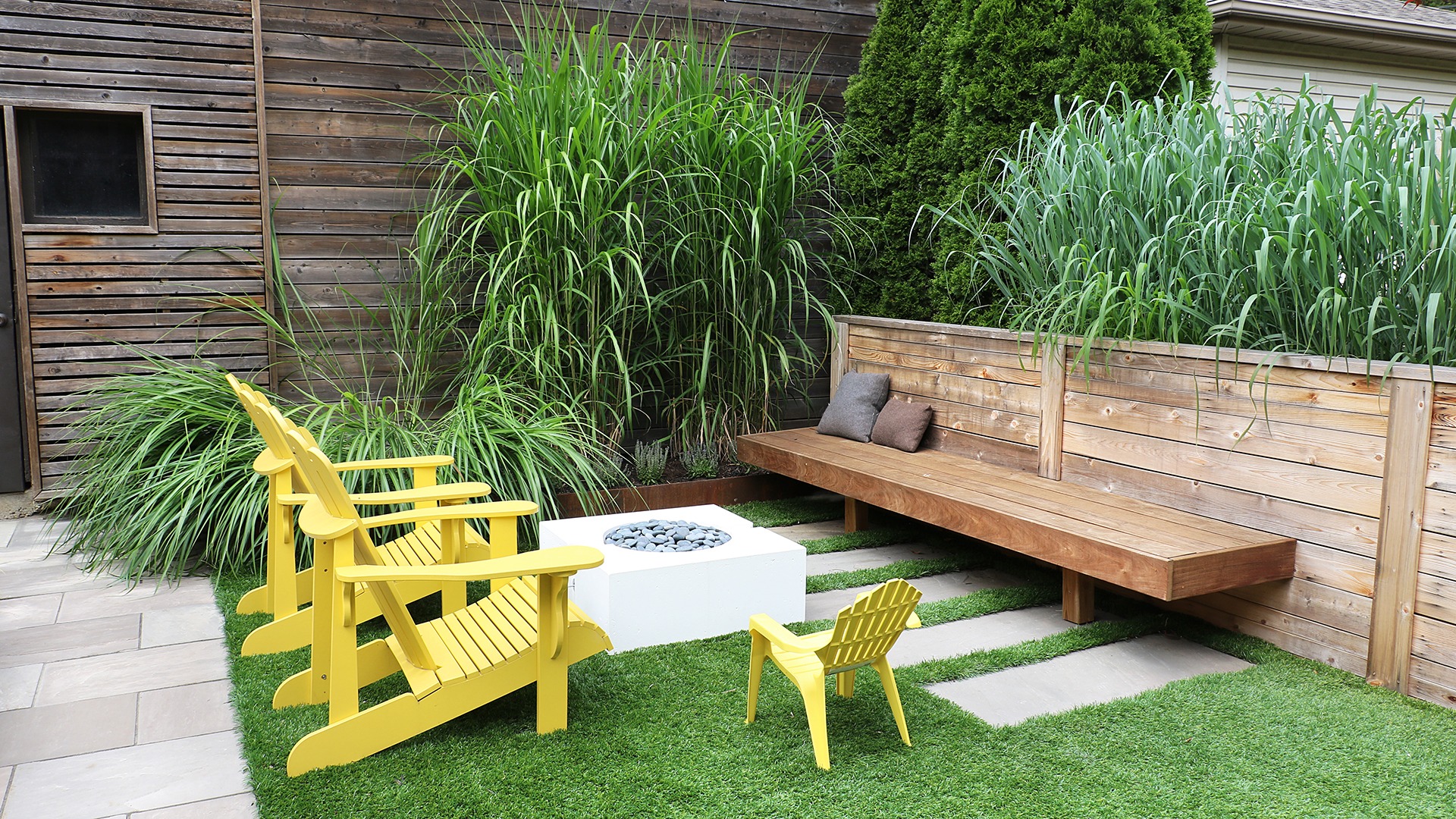 Cozy garden setting with wooden bench, yellow chairs, and fire pit. Surrounded by tall grass, wooden fence, and patio stones.