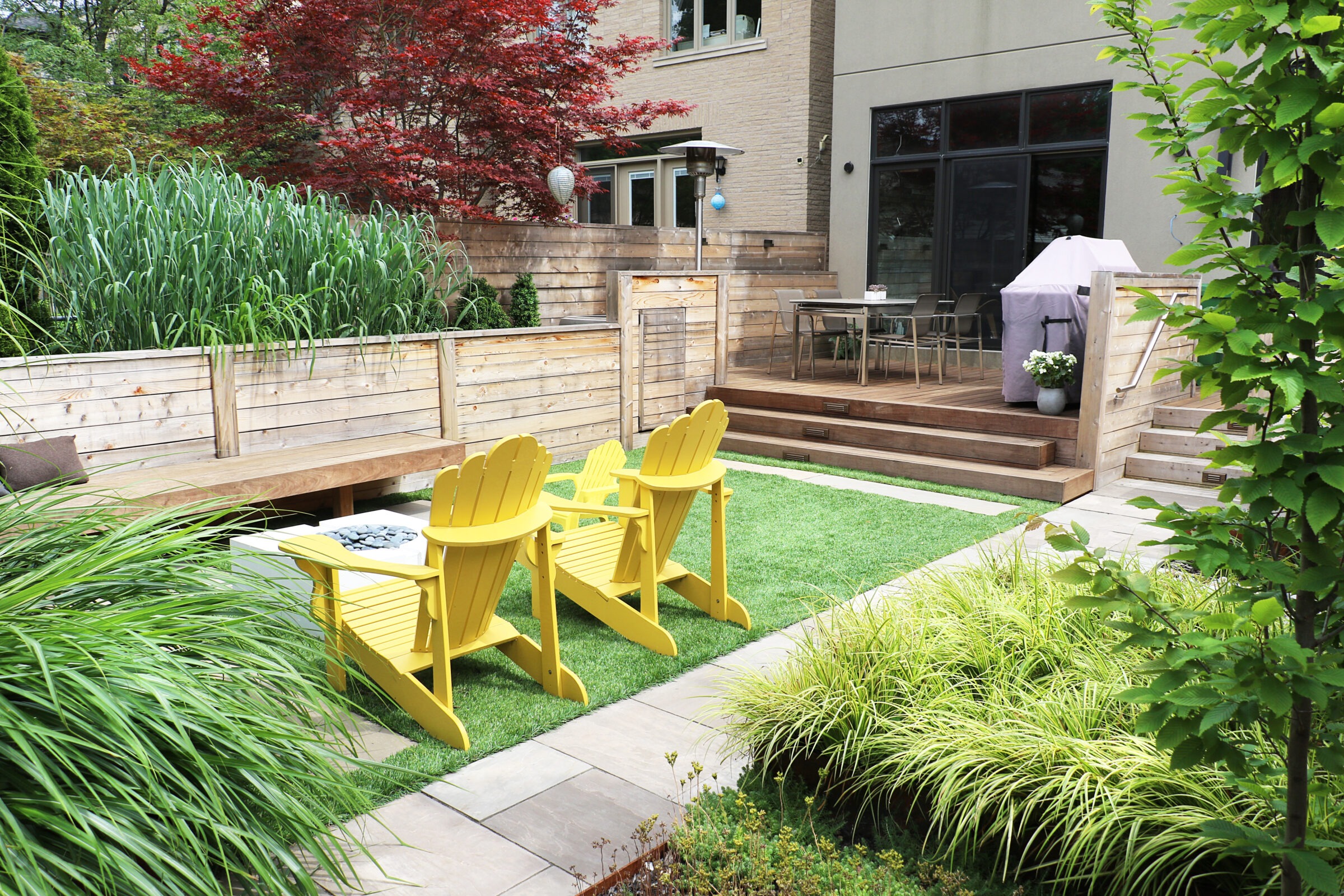 A modern backyard features yellow chairs, wooden fencing, lush green plants, and a wooden deck with outdoor furniture and a covered grill.