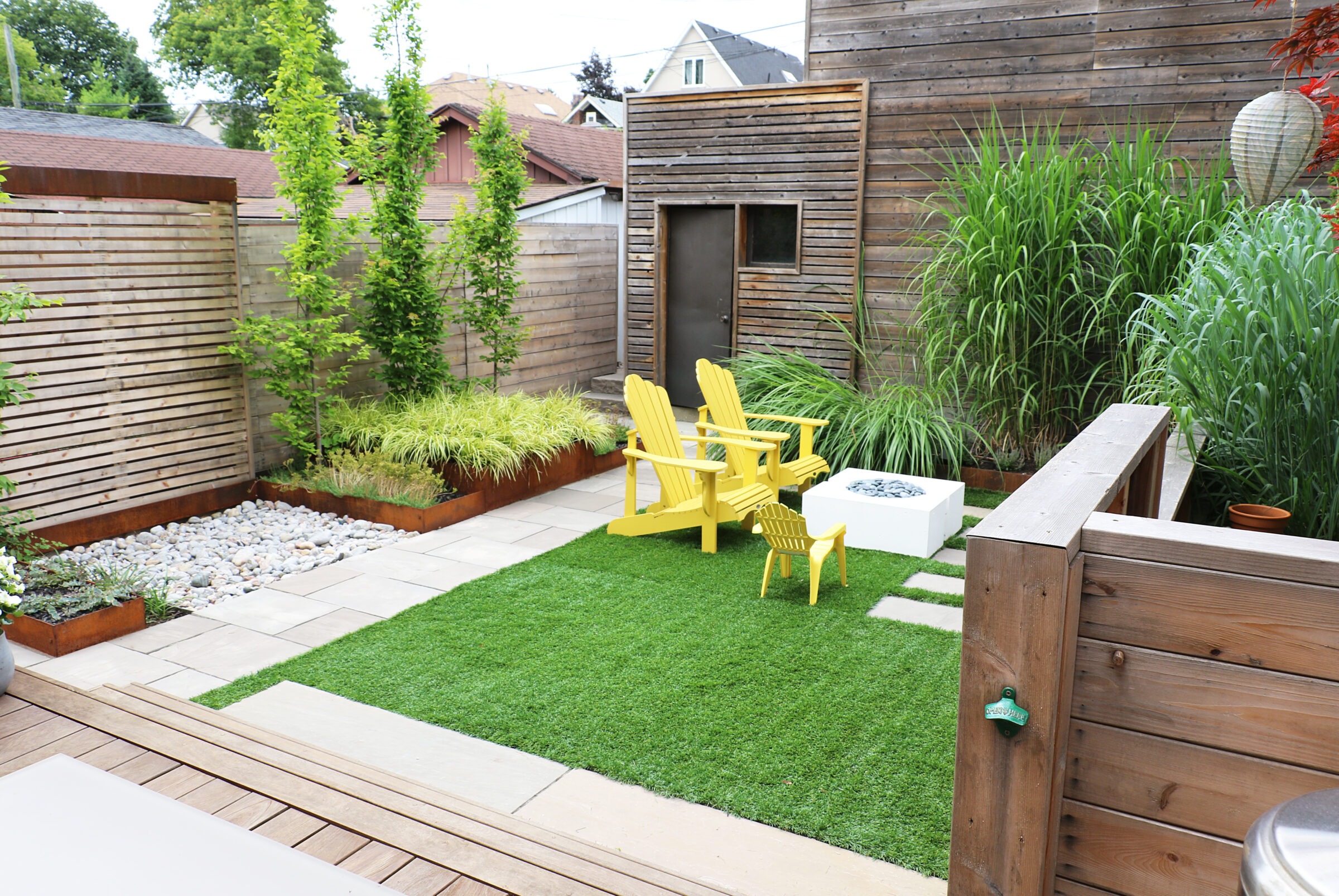 Modern backyard with wooden fencing, lush greenery, yellow chairs on artificial grass, stone pathway, decking, and a white fire pit. Cozy, tranquil atmosphere.