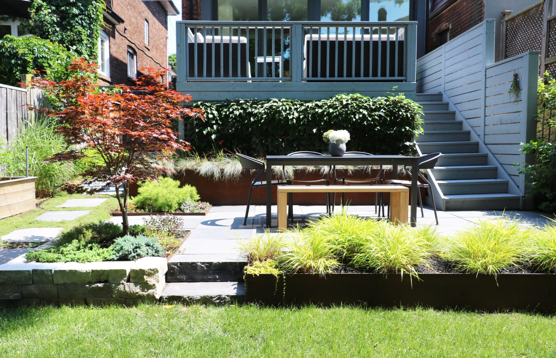 A modern backyard patio with a dining table, chair, vibrant plants, and a staircase leading to a house, surrounded by lush greenery.