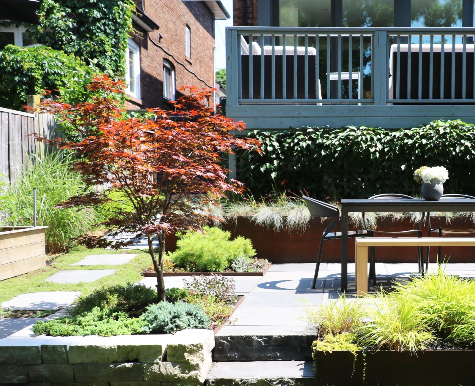 A modern backyard patio with a dining table, chair, vibrant plants, and a staircase leading to a house, surrounded by lush greenery.