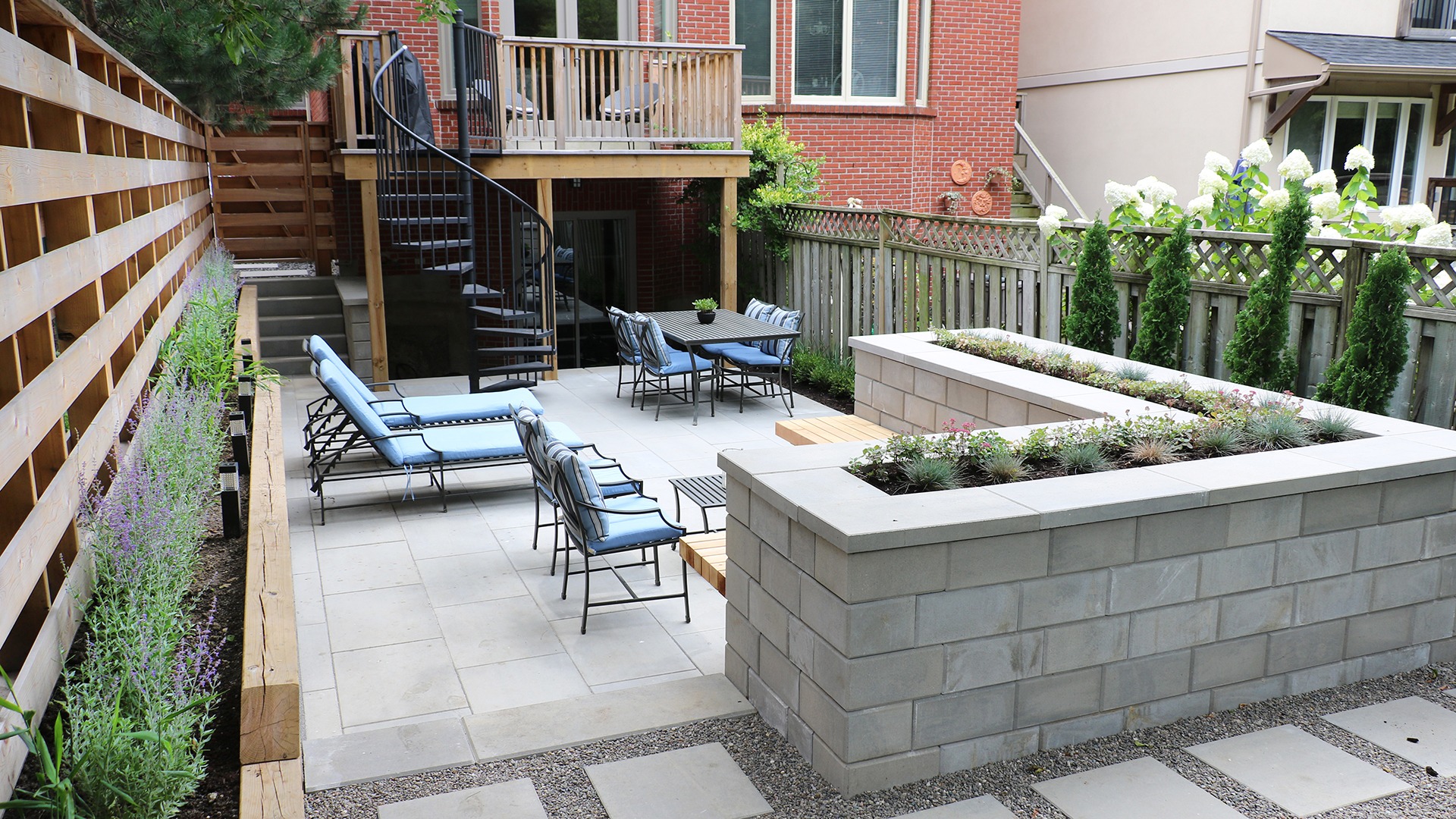 Modern backyard patio with lounge chairs, dining set, and spiral staircase. Surrounded by wooden fencing, plants, and brick walls. Cozy outdoor space.