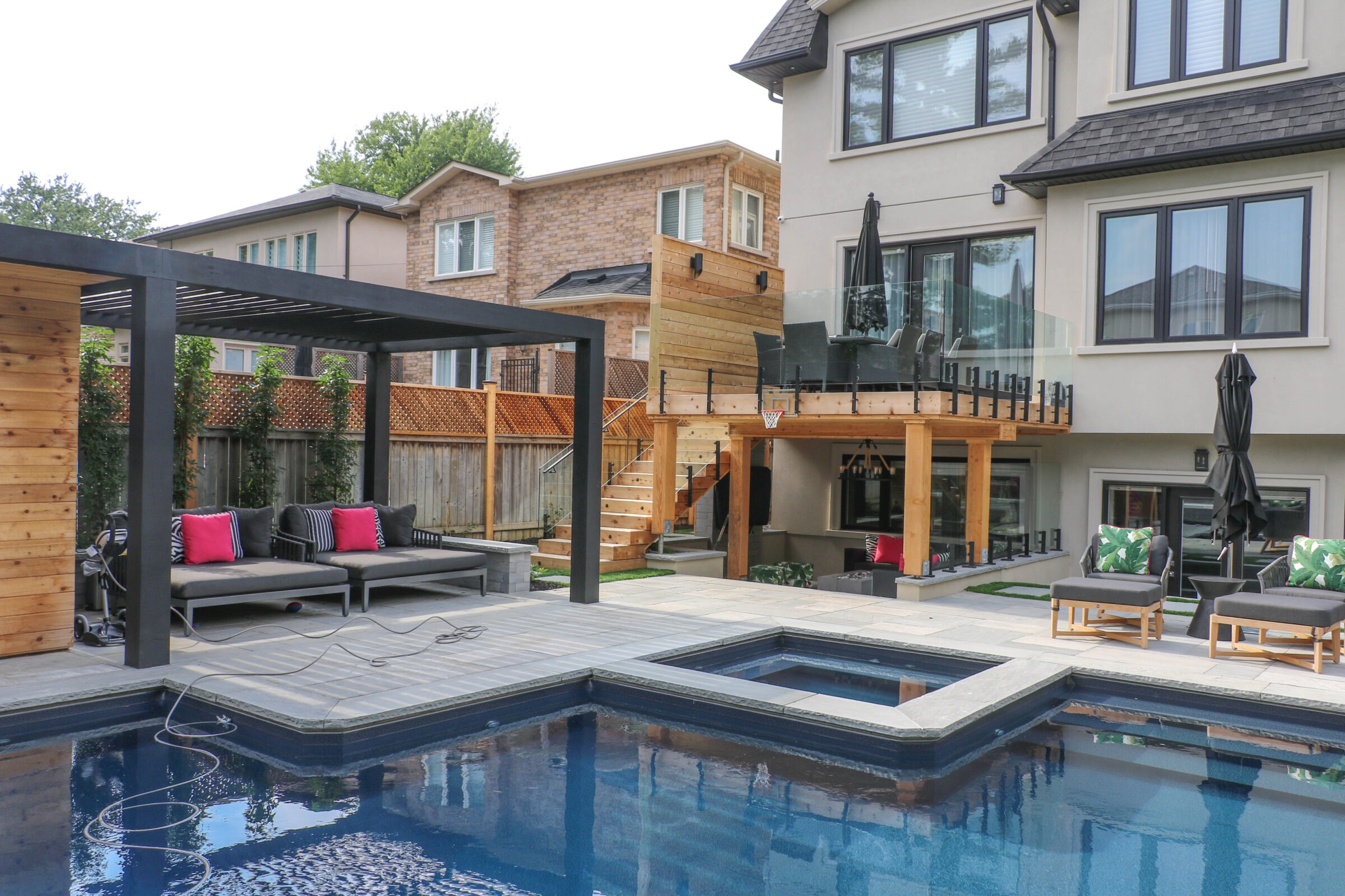 A modern backyard with a swimming pool, lounge chairs, pergola, and patio. Contemporary house with wooden accents and glass railings behind.