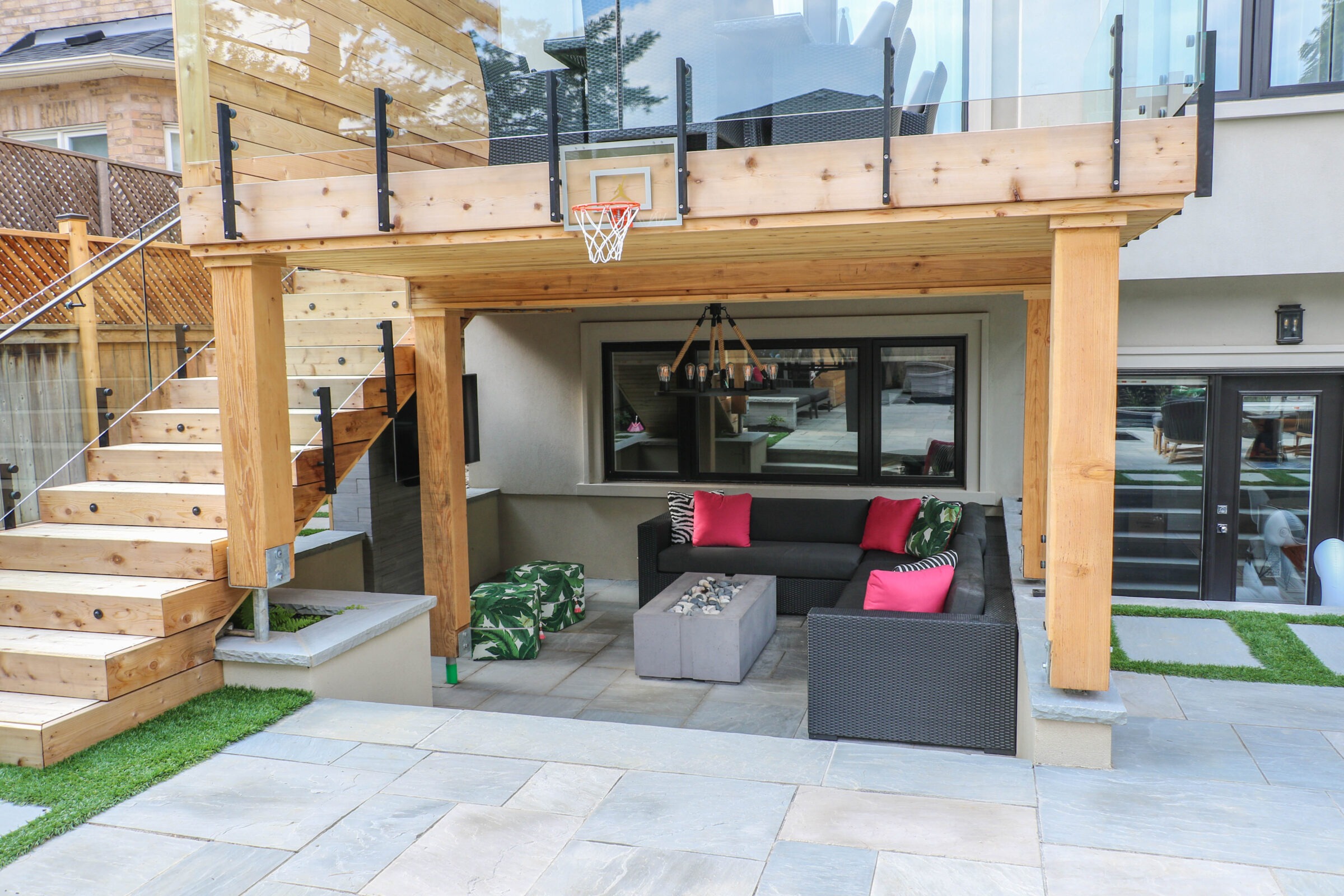 Modern backyard patio with wooden staircase, glass railing, and basketball hoop. Cozy seating area with bright cushions and outdoor lighting.