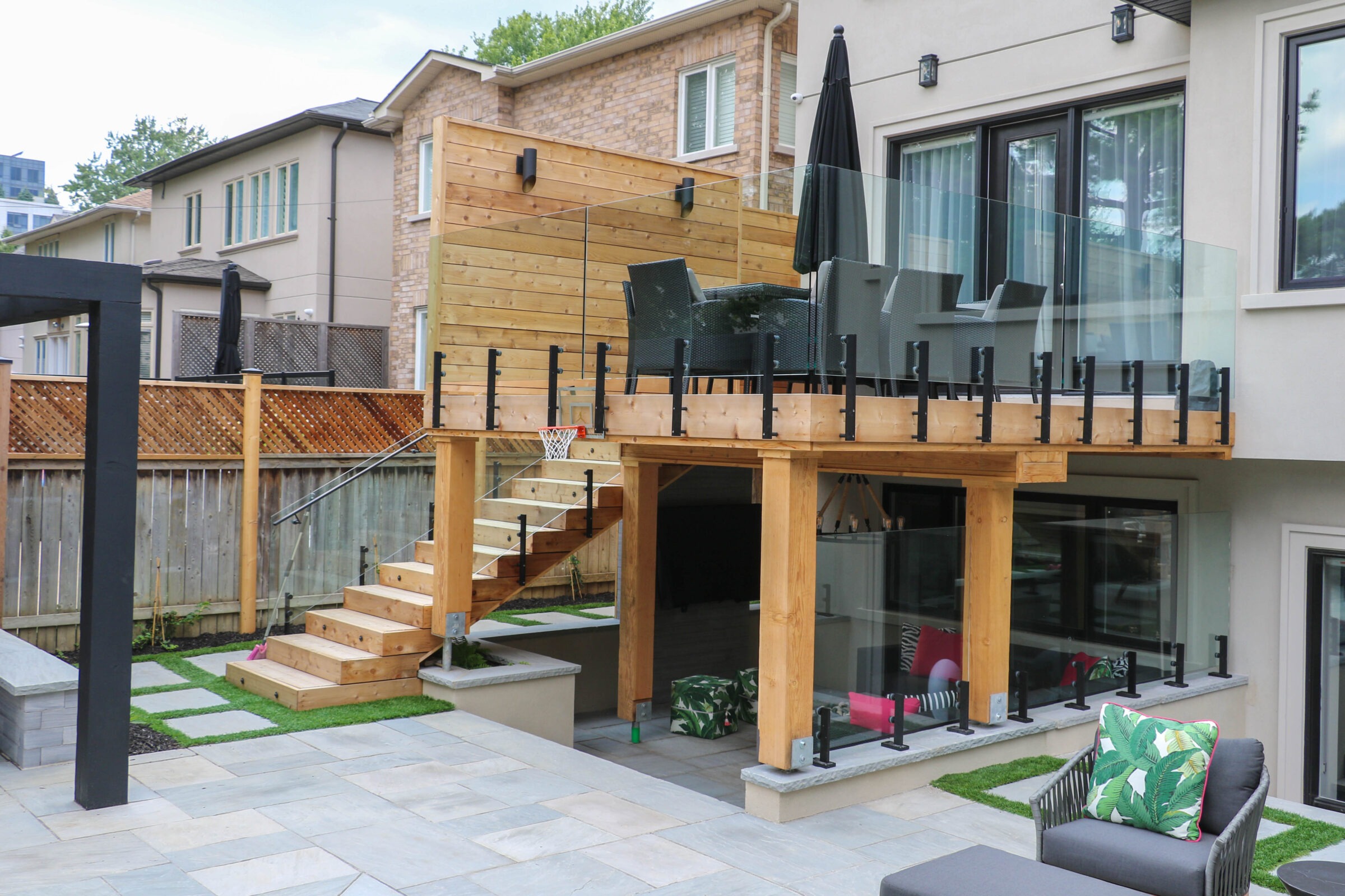 Modern backyard patio with wooden deck, outdoor furniture, glass railing, and stairs. Cozy seating area underneath with patterned cushions and greenery.