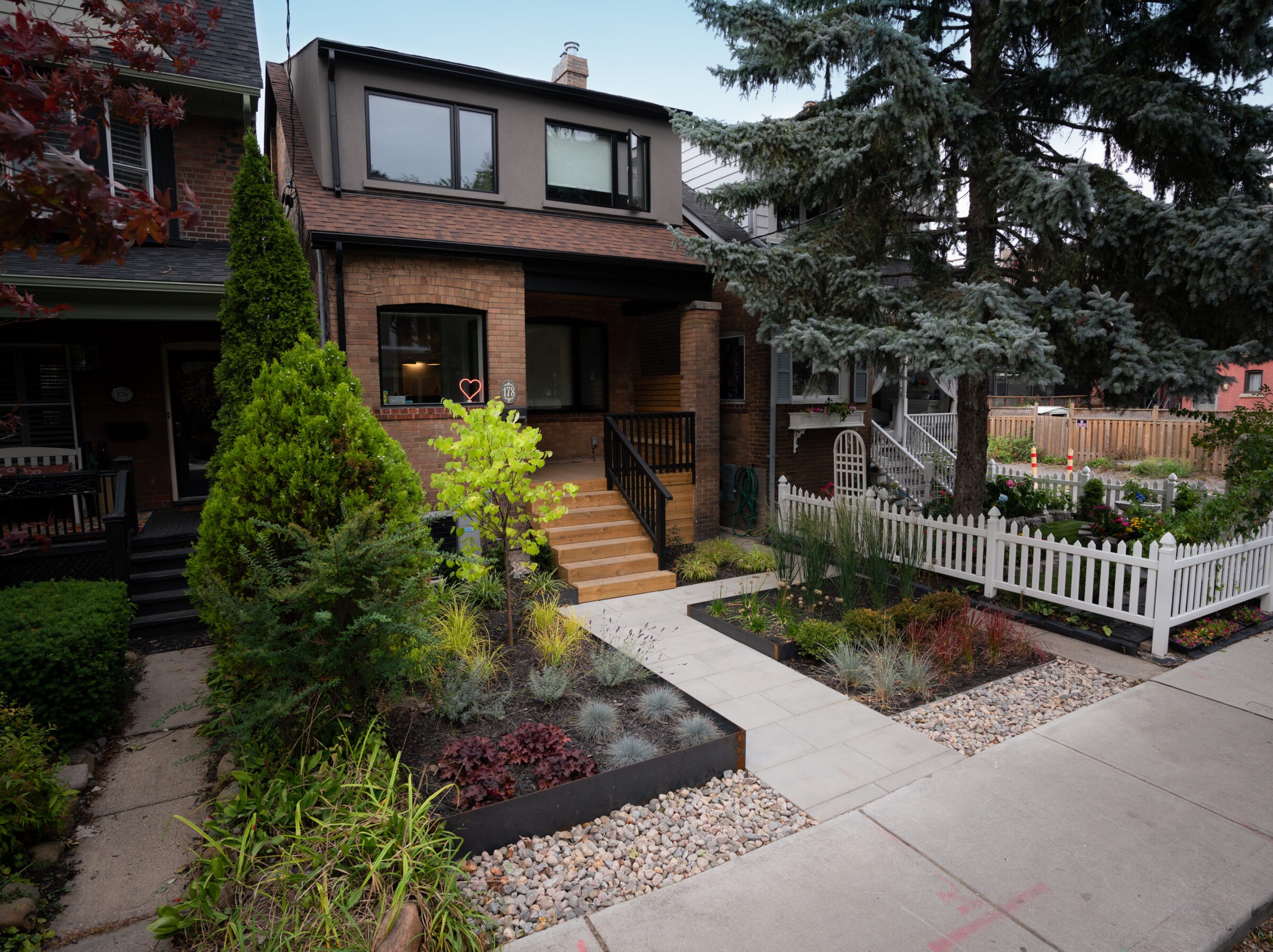 A cozy brick house with a manicured garden, white picket fence, and pathway. Lush greenery and trees frame the scene.