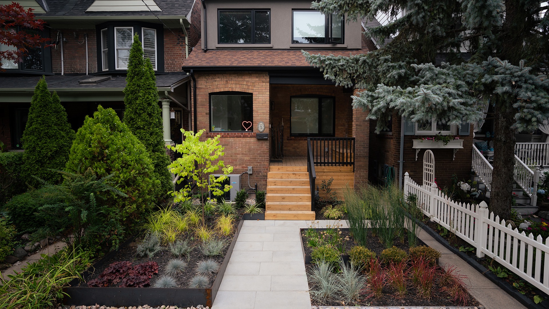 Charming brick house with landscaped front yard, including diverse plants and wooden steps, accompanied by neighboring homes and a white picket fence.