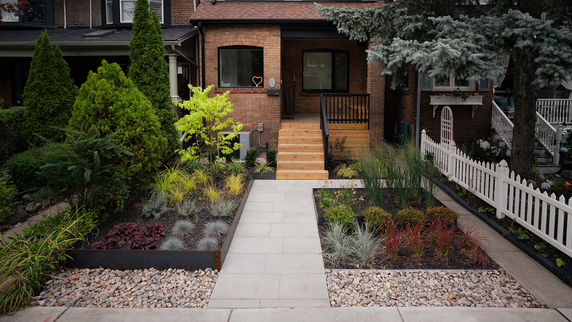 A neatly landscaped front yard with colorful plants, small trees, and a brick house featuring a porch and a white picket fence.