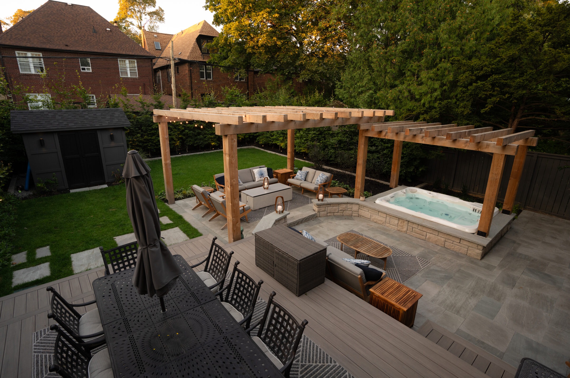 A backyard with a pergola, hot tub, and outdoor seating surrounded by greenery and brick houses, providing a cozy relaxation space.