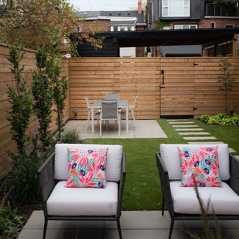 A cozy backyard features a patio with chairs, colorful cushions, a wooden fence, and dining area, surrounded by greenery and urban buildings.