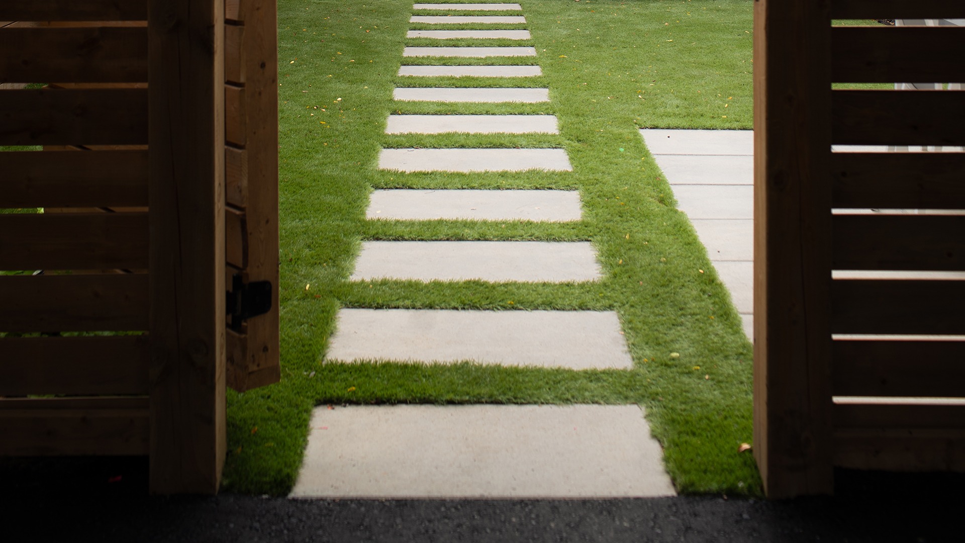 A neatly landscaped garden path with rectangular stepping stones leads through lush green grass, viewed from an open wooden gate on a cloudy day.