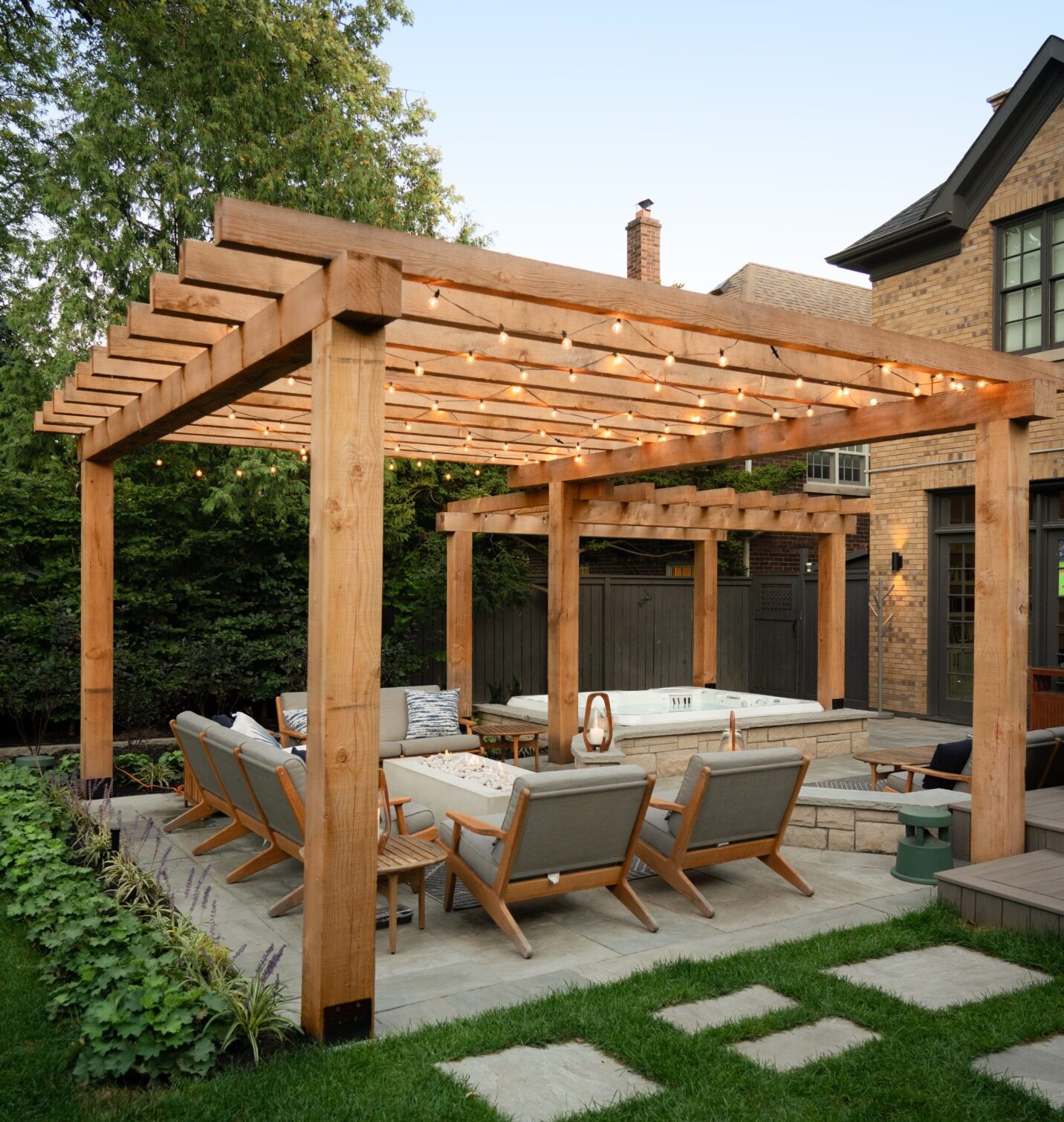 A backyard with a wooden pergola, string lights, cozy seating, and a fire pit, adjacent to a brick house amidst lush green surroundings.