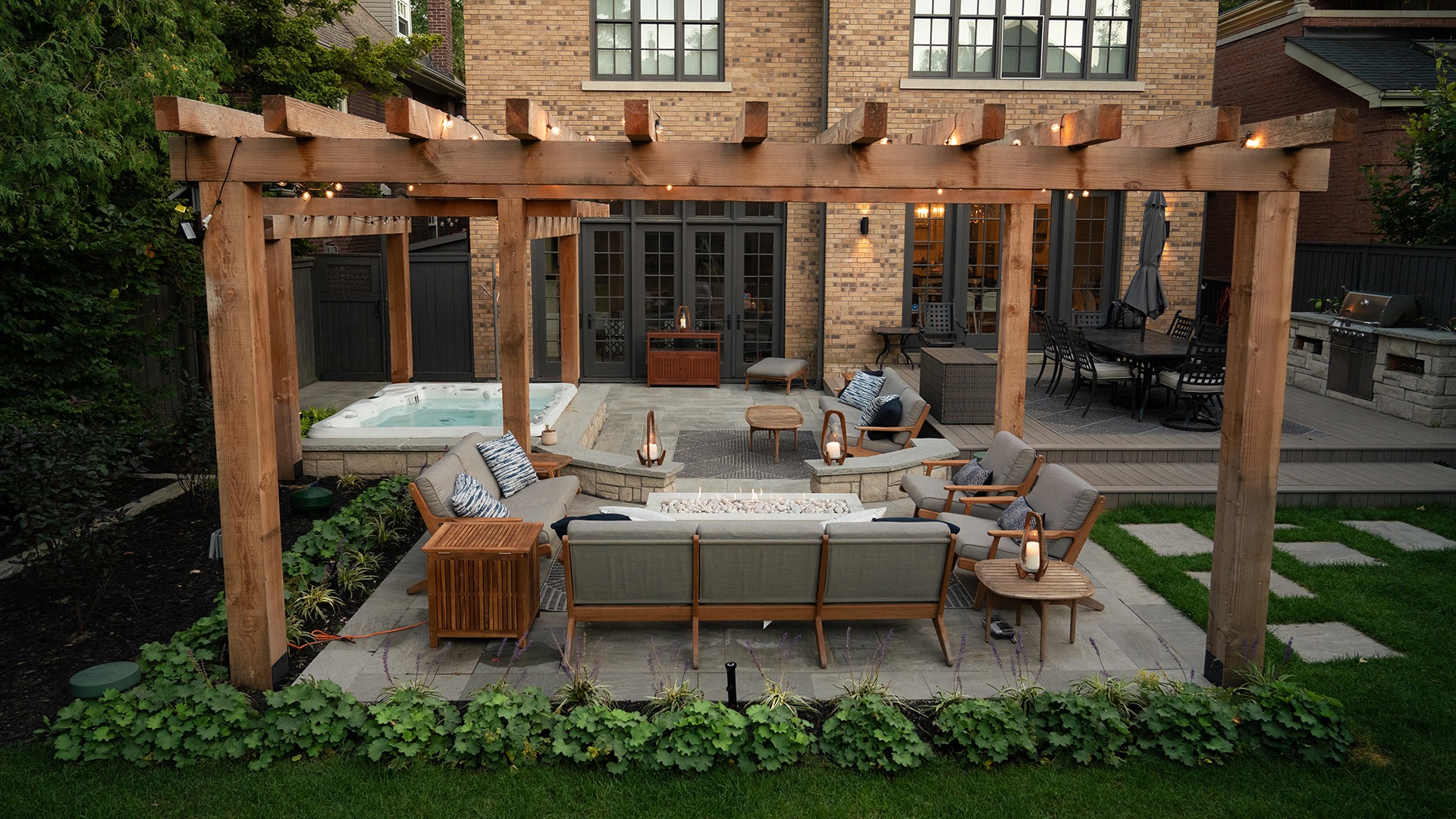 Modern backyard with cozy seating under a wooden pergola, hot tub, and grill area, adjacent to a brick house with large windows.
