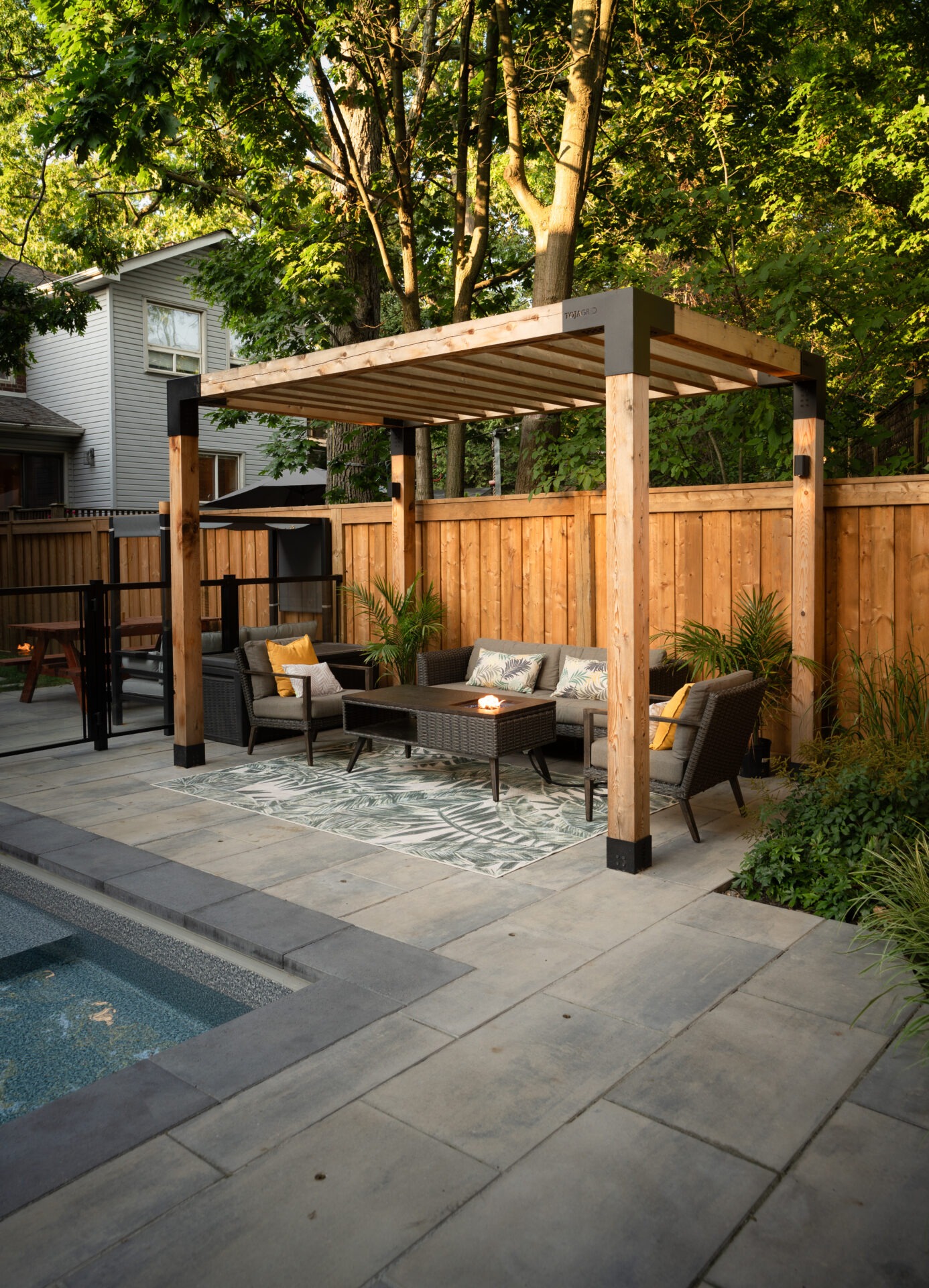A cozy backyard patio with a pergola, comfortable seating, decorative rug, and plants, beside a small pool surrounded by trees and a fence.