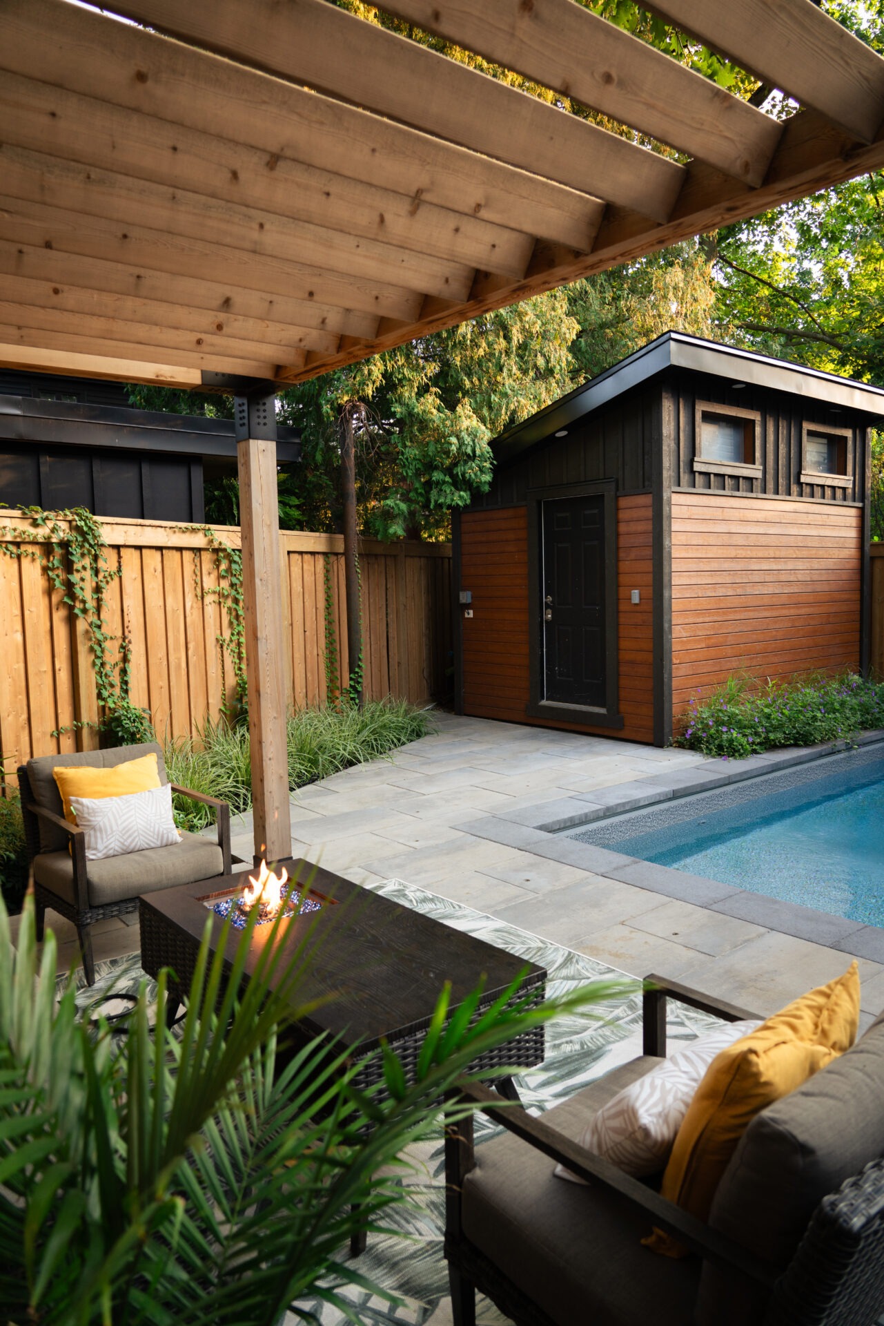 A backyard patio with a pool, wooden pergola, fire pit, and cozy seating area beside a well-maintained garden and small shed.