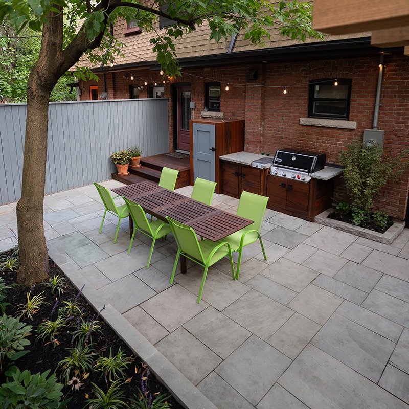 A cozy backyard patio features a wooden dining table with green chairs, grill, potted plants, and string lights against a brick house.
