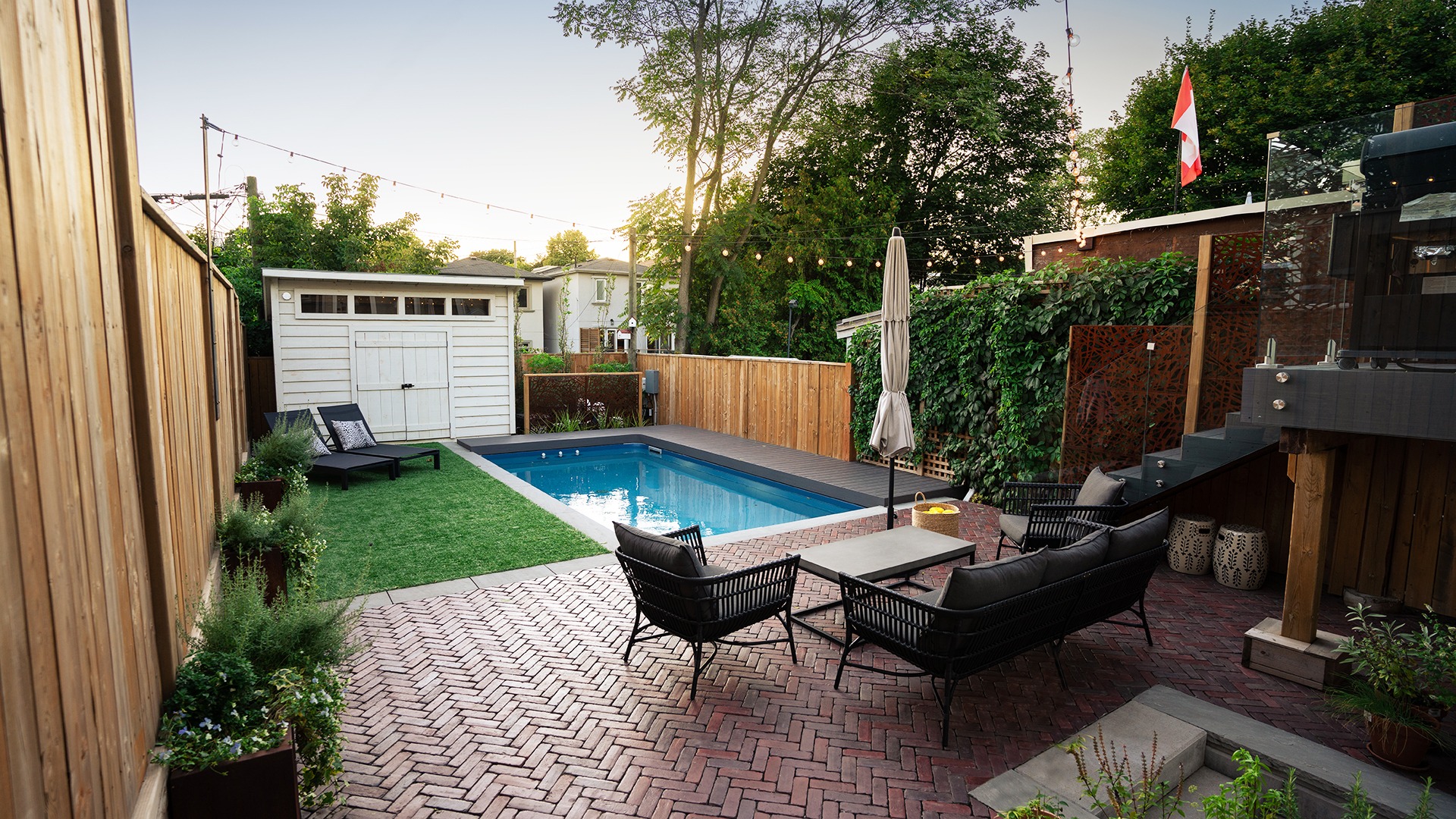Backyard features a small swimming pool, wooden deck, brick patio, and lounge chairs. Surrounded by fences and greenery under the evening sky.