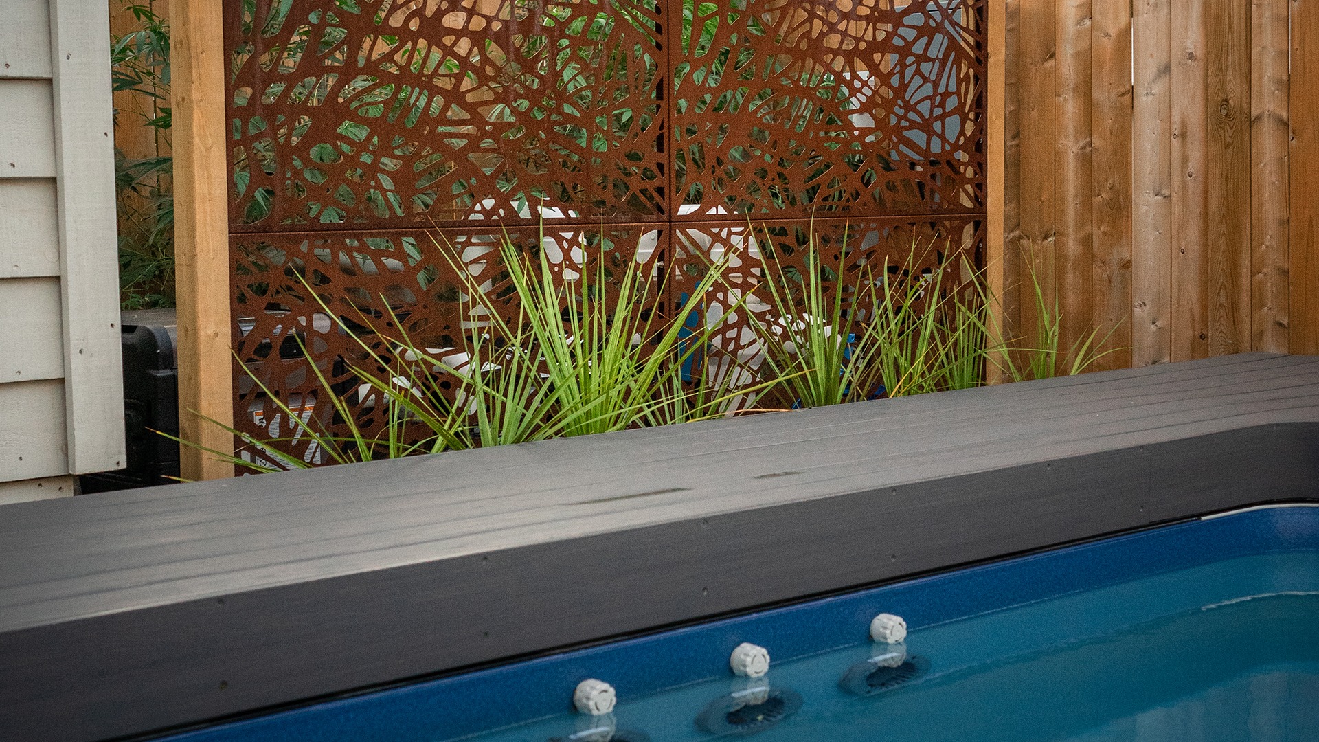 Outdoor space featuring a modern privacy screen with geometric patterns, wooden fencing, green plants, and the edge of a blue hot tub.