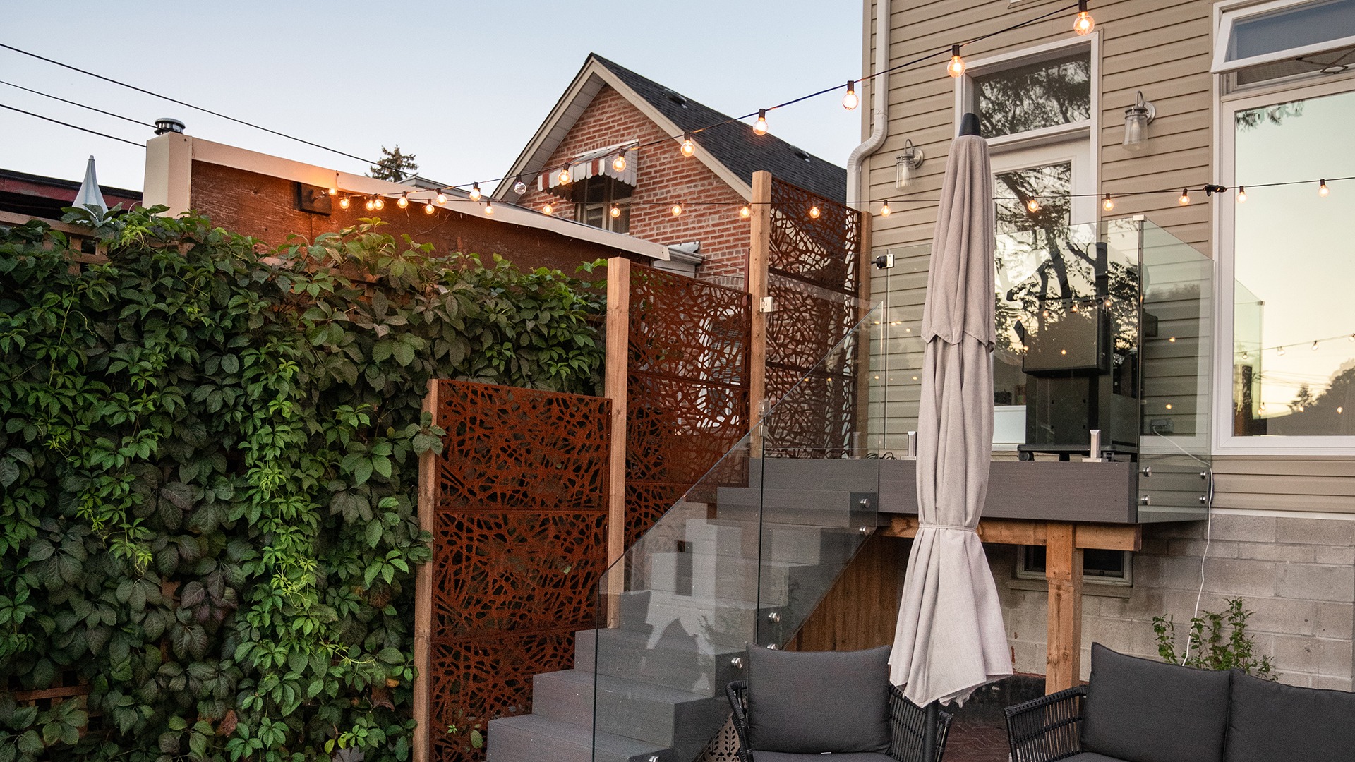 Modern patio with wooden steps, decorative panels, string lights, and an umbrella. Lush green vines cover the wall, creating a cozy atmosphere.
