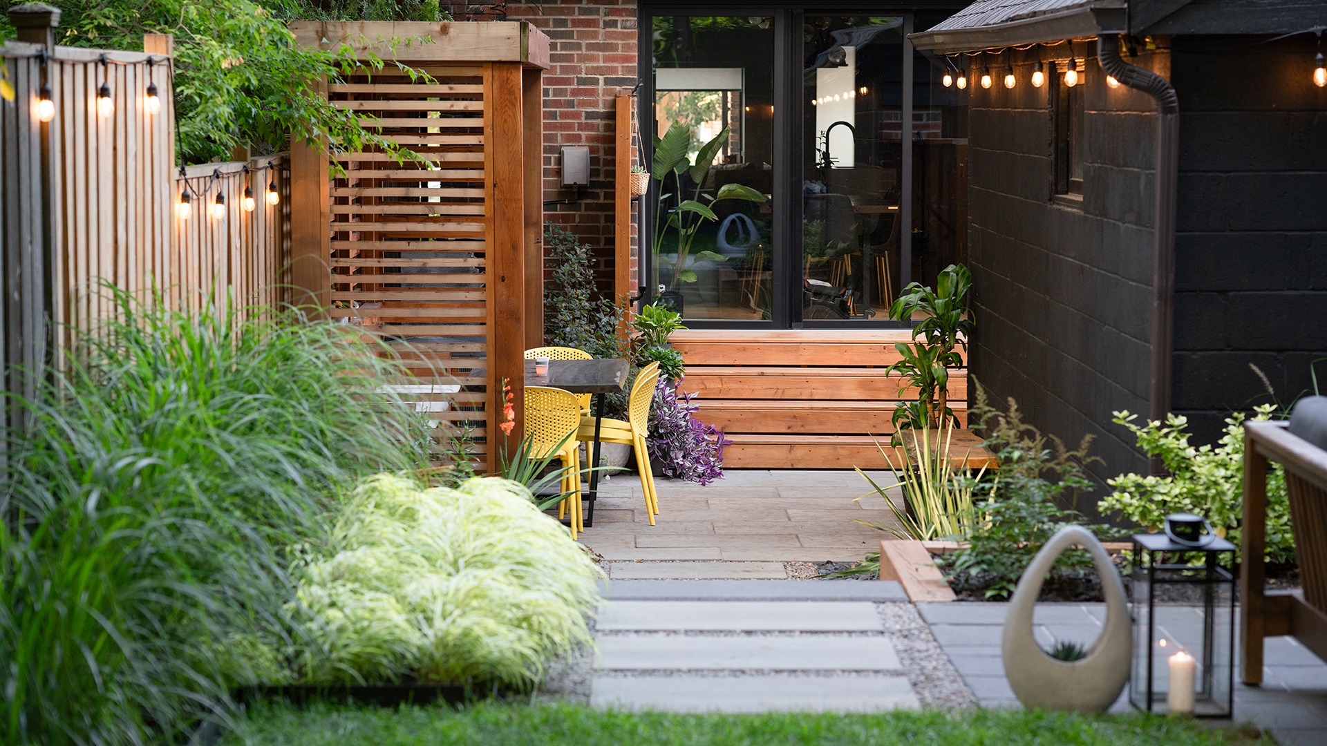 Cozy backyard patio with wooden fencing, string lights, and lush plants. Features yellow chairs, a small table, and a modern wooden bench.