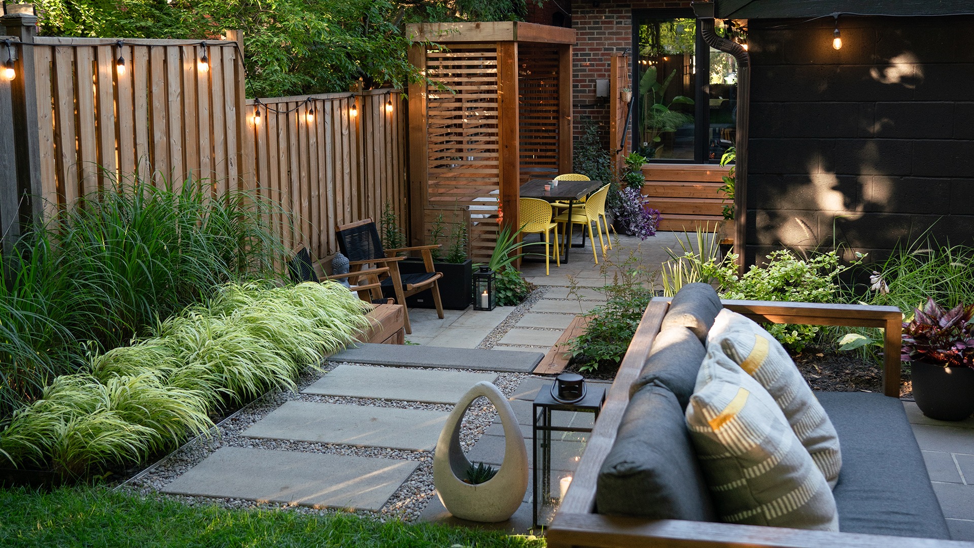 Modern backyard featuring a cozy seating area, string lights, lush greenery, and a wooden fence, creating a serene outdoor space for relaxation.