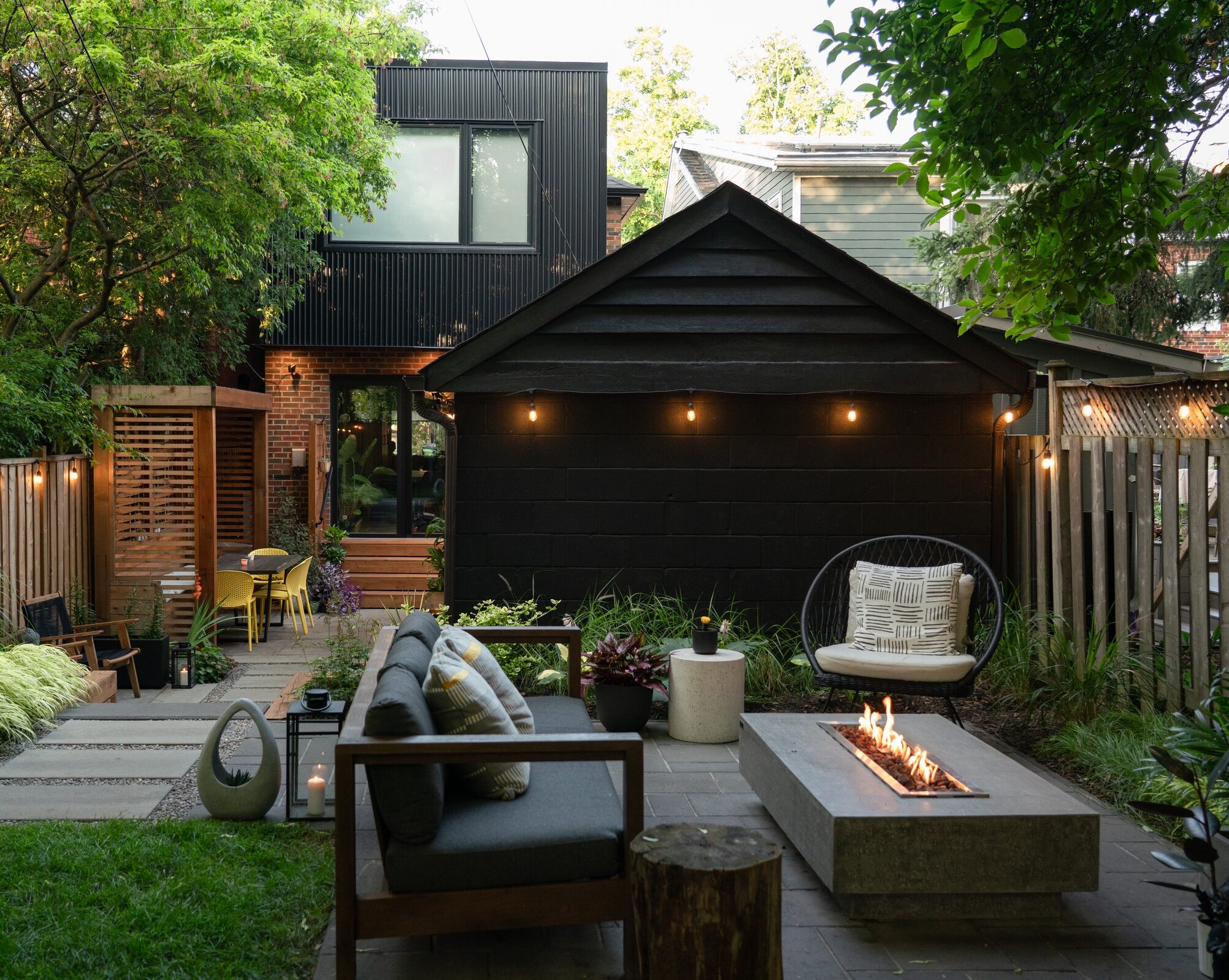 Modern backyard with a cozy seating area, sleek fire pit, garden, and dining table. Surrounded by lush greenery and contemporary decor.