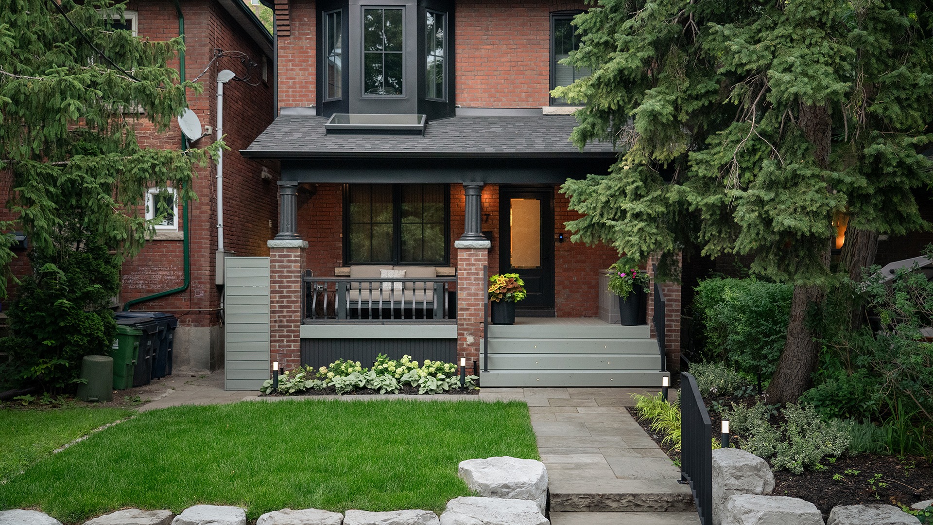 A charming brick house with porch, surrounded by lush greenery and trees, features a neatly maintained lawn and potted plants.
