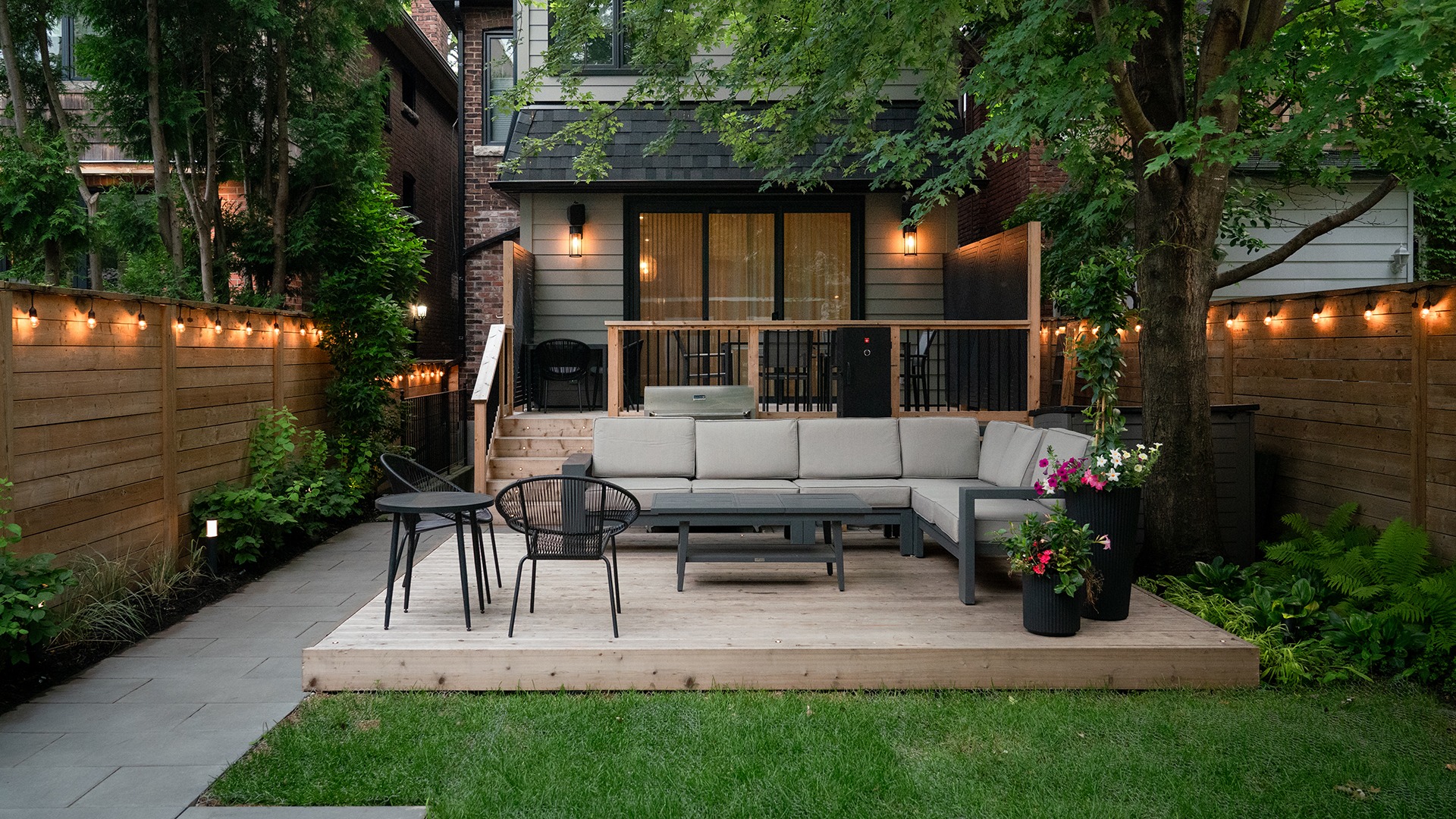 Cozy backyard with wooden deck, outdoor seating, string lights, and lush greenery surrounding a modern house during the evening.