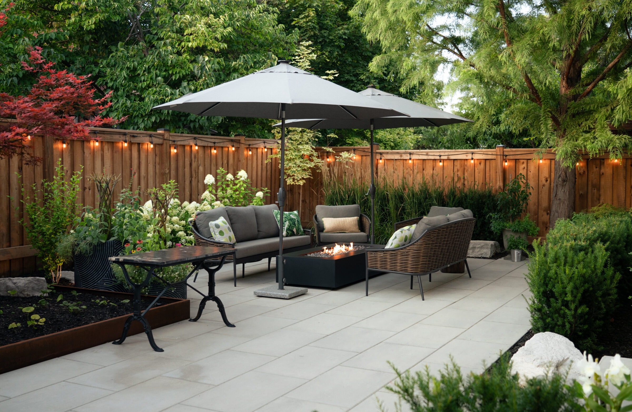 A cozy, modern patio features wicker furniture, cushions, a fire pit, umbrellas, surrounded by lush greenery and soft string lights on a wooden fence.