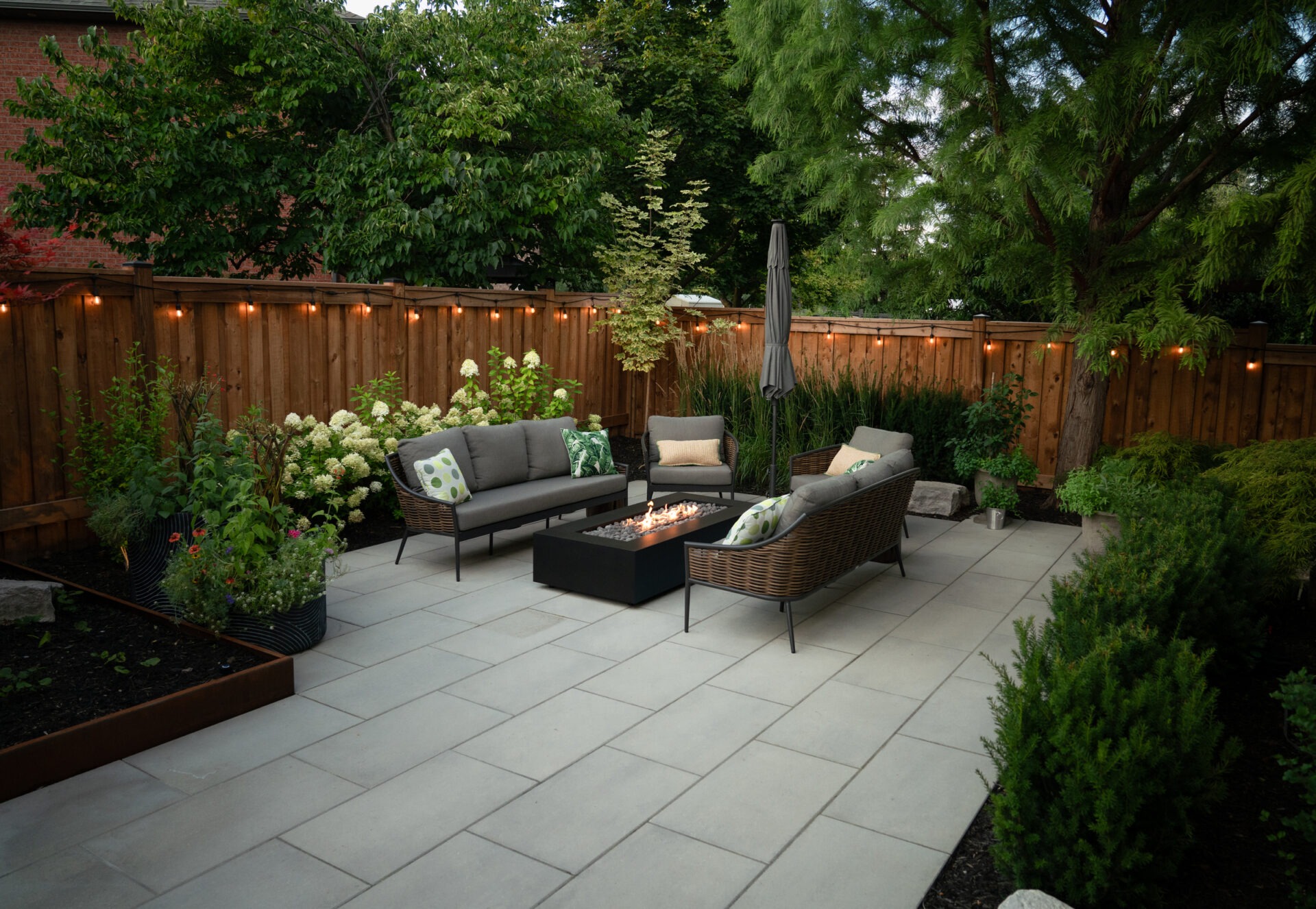 A cozy backyard patio features modern furniture, a central fire pit, string lights, and lush greenery enclosed by a wooden fence.