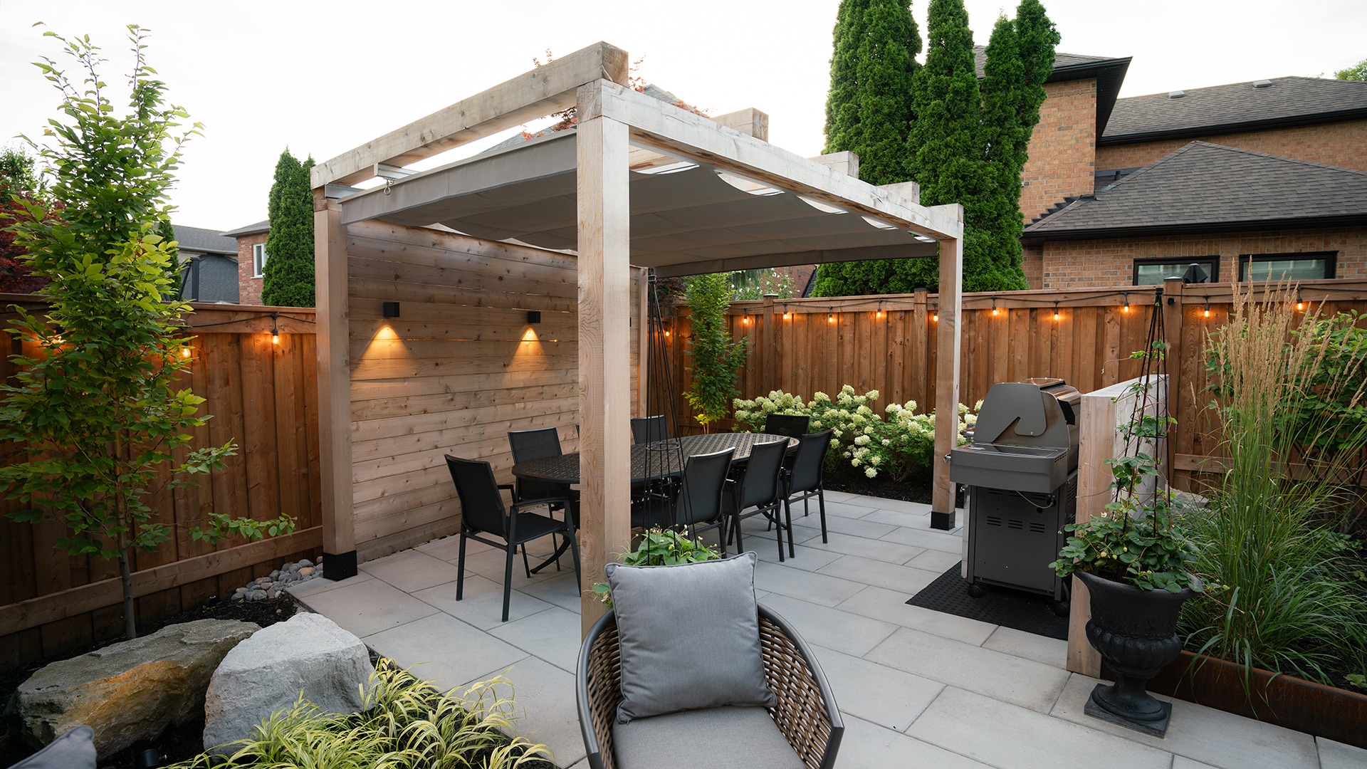 Cozy backyard patio with a pergola, dining table, grill, plants, and warm string lights on wooden fence, creating an inviting outdoor space.