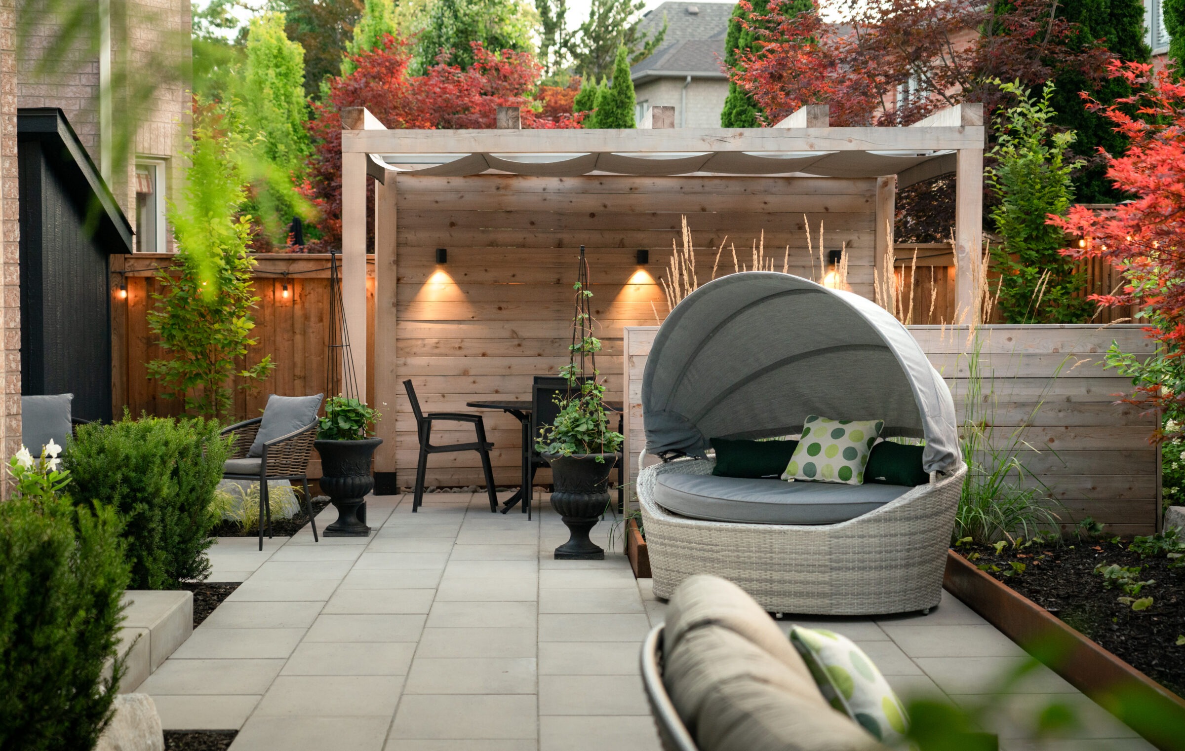 A cozy backyard patio features a wooden pergola, cushioned seating, ambient lighting, and lush greenery, creating an inviting outdoor relaxation space.