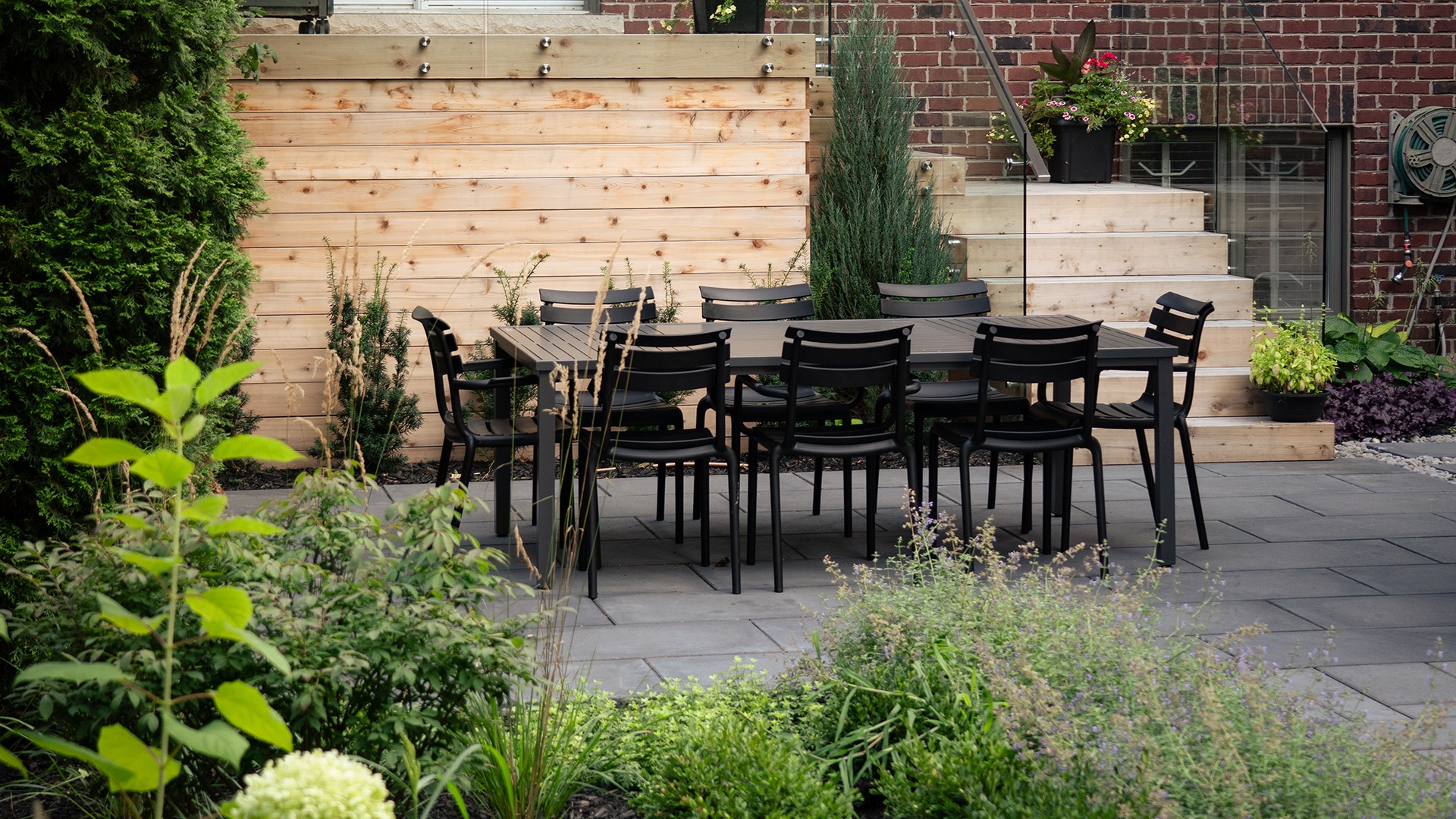 A cozy patio features a black dining set surrounded by lush green plants, wooden accents, and brick walls, creating an inviting outdoor space.