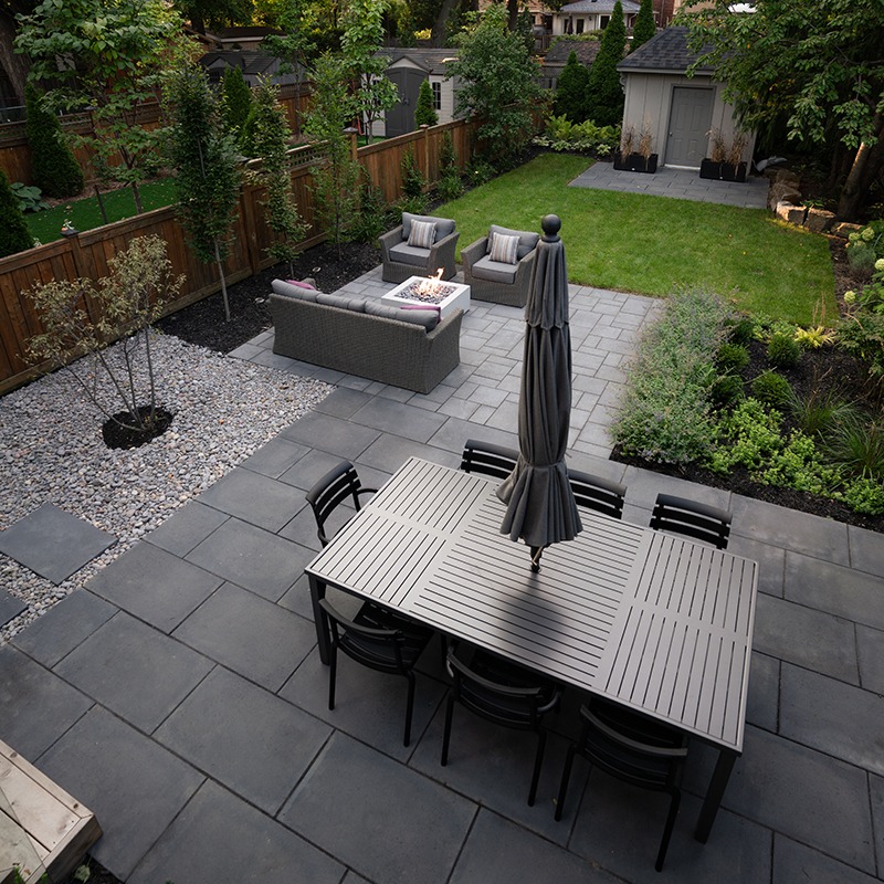 Modern backyard with patio dining set, fire pit, and lush garden. Stone tiles and wooden fence create a cozy, inviting outdoor space.