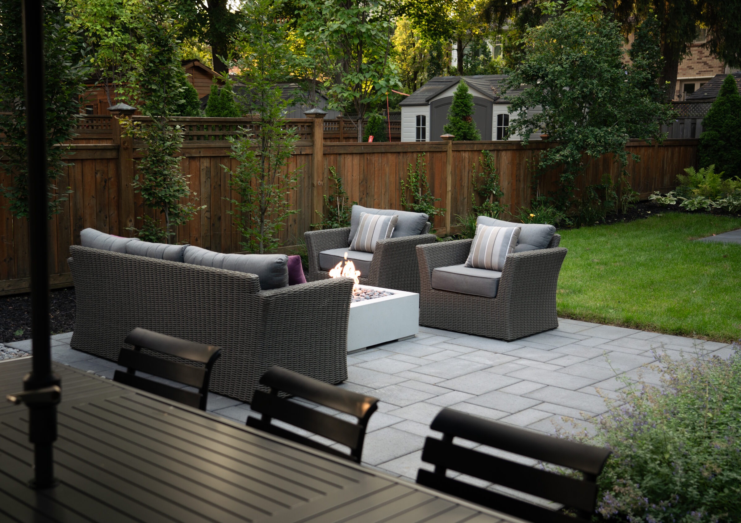 A cozy backyard patio features wicker furniture around a lit fire pit, with lush greenery and wooden fencing creating a tranquil outdoor space.