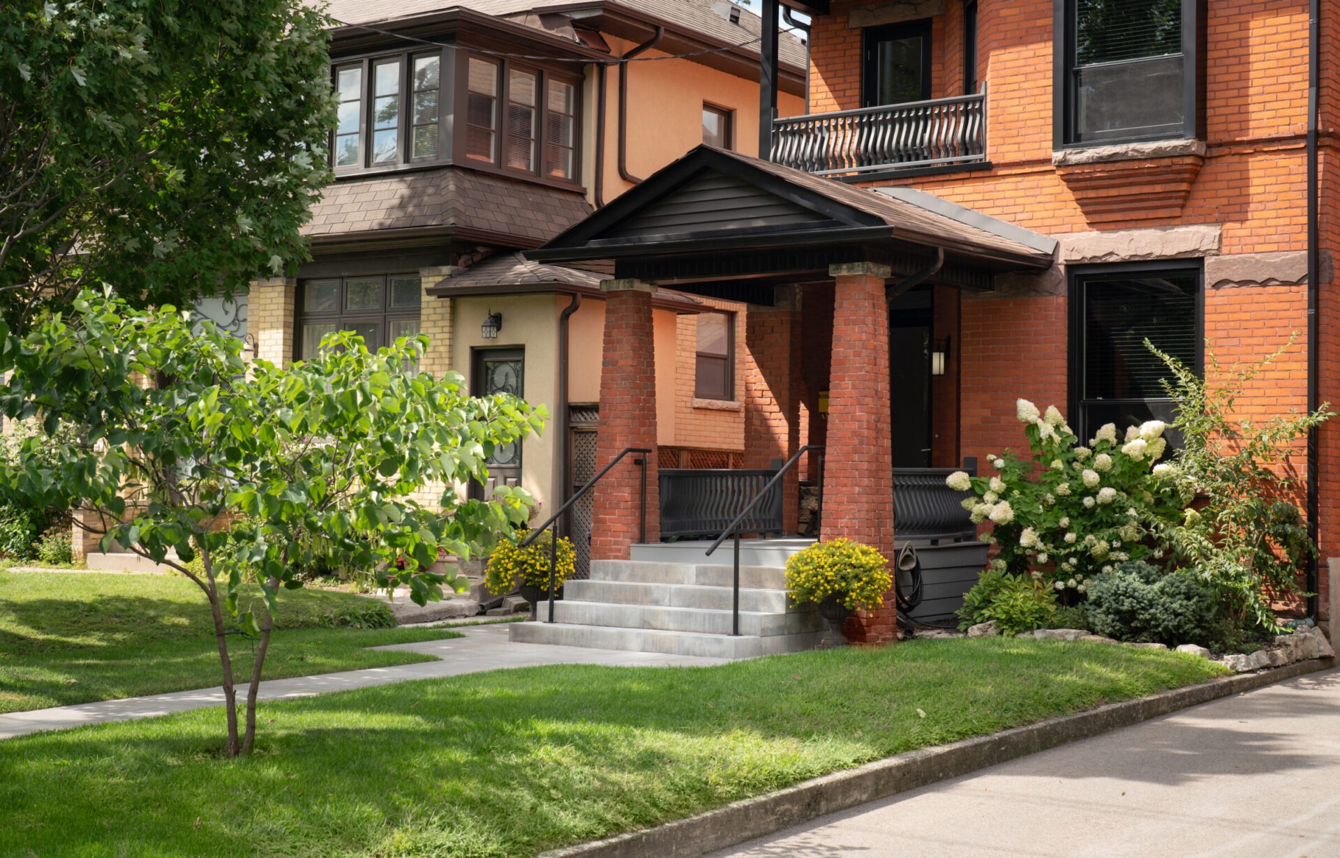 Red-brick house with porch, surrounded by lush green garden and small tree. Adjacent houses feature unique architecture and complement the tranquil suburban setting.