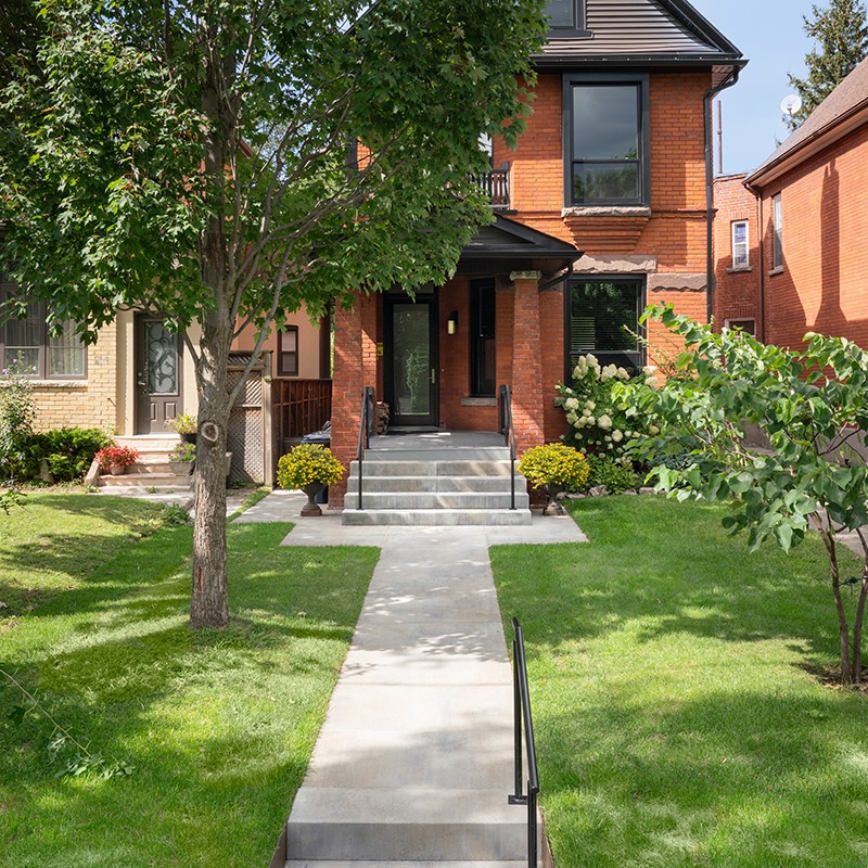Brick house with black accents, surrounded by trees and bushes. Concrete pathway leads to entrance, flanked by stairs. Peaceful residential neighborhood setting.