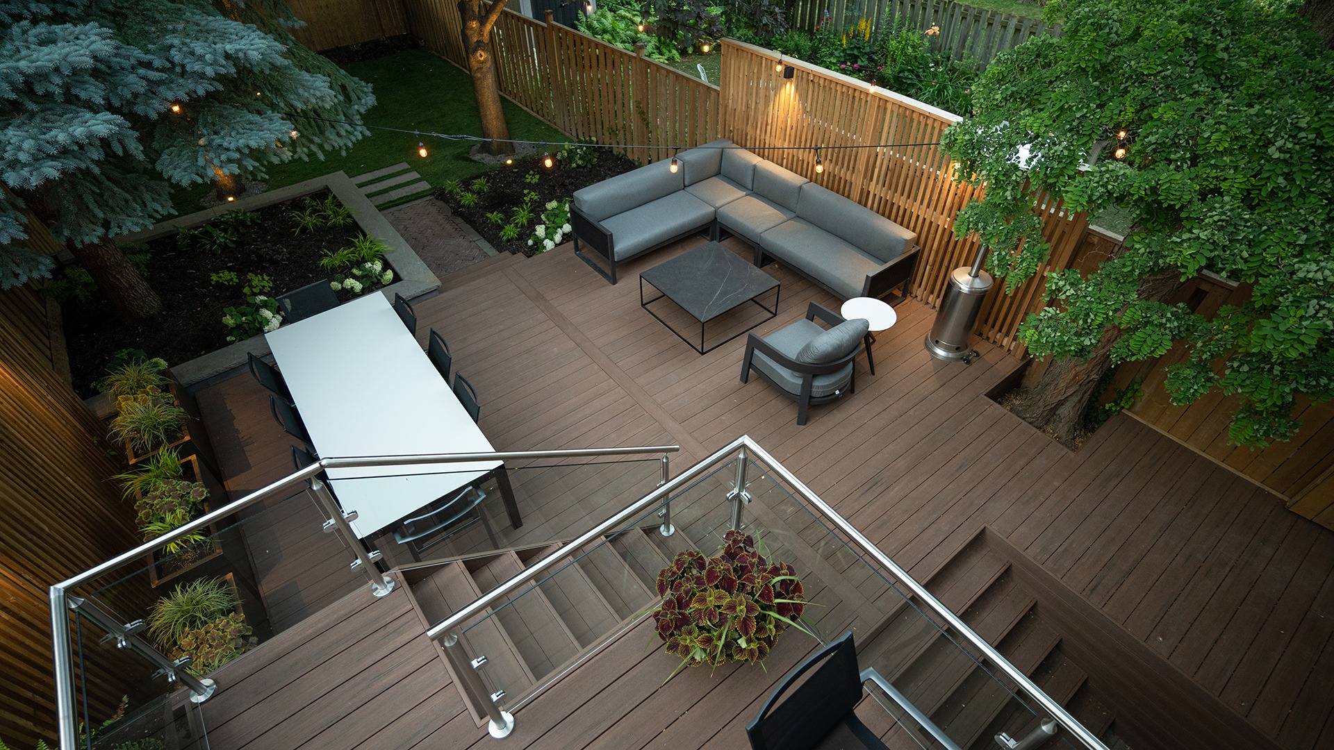 Modern backyard patio with wooden decking, sectional sofa, dining table, and string lights. Surrounded by greenery and trees for a cozy atmosphere.