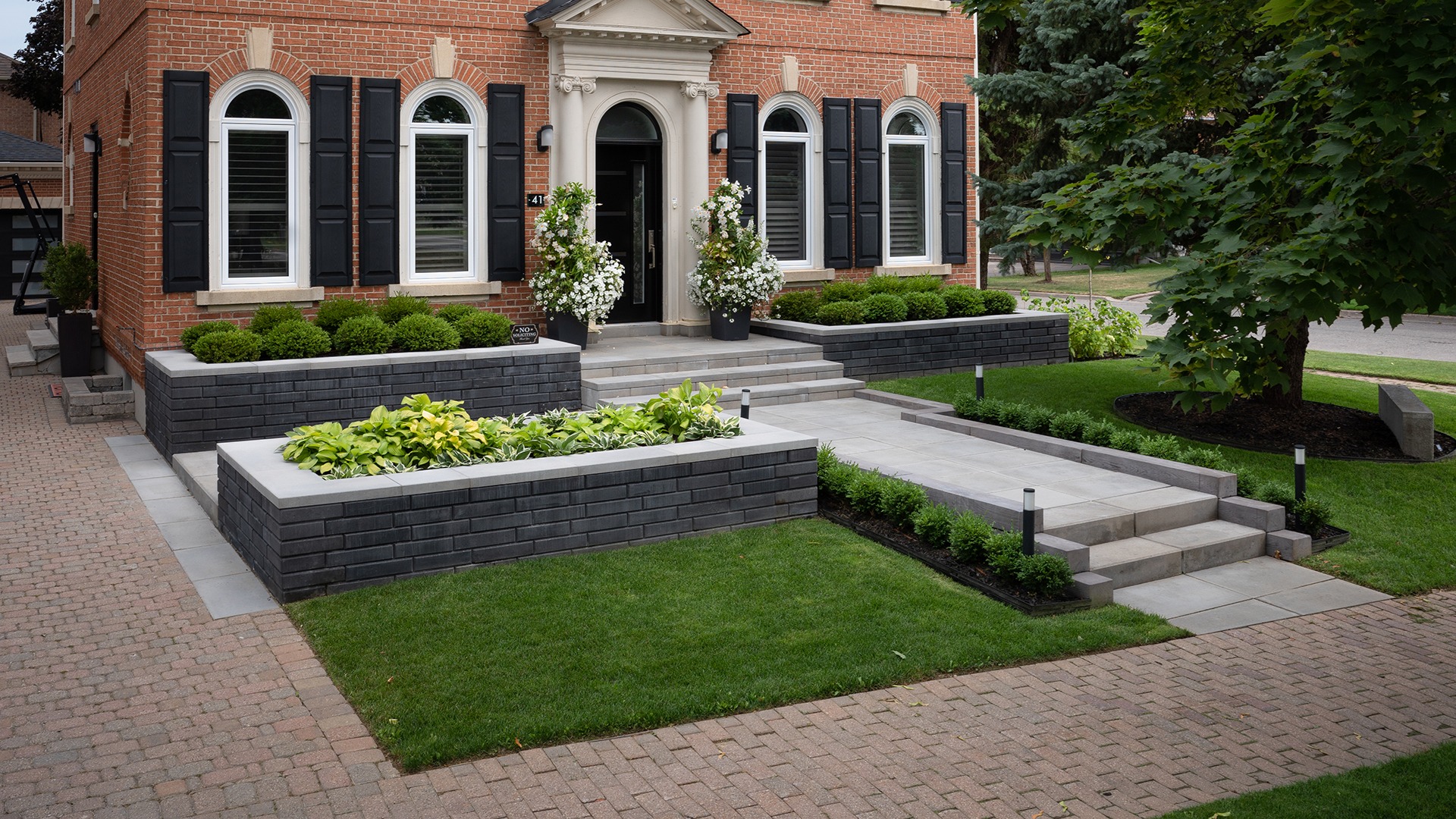 A red brick house features black shutters, arched windows, and manicured landscaping with plants and pathways, creating a welcoming front entrance.