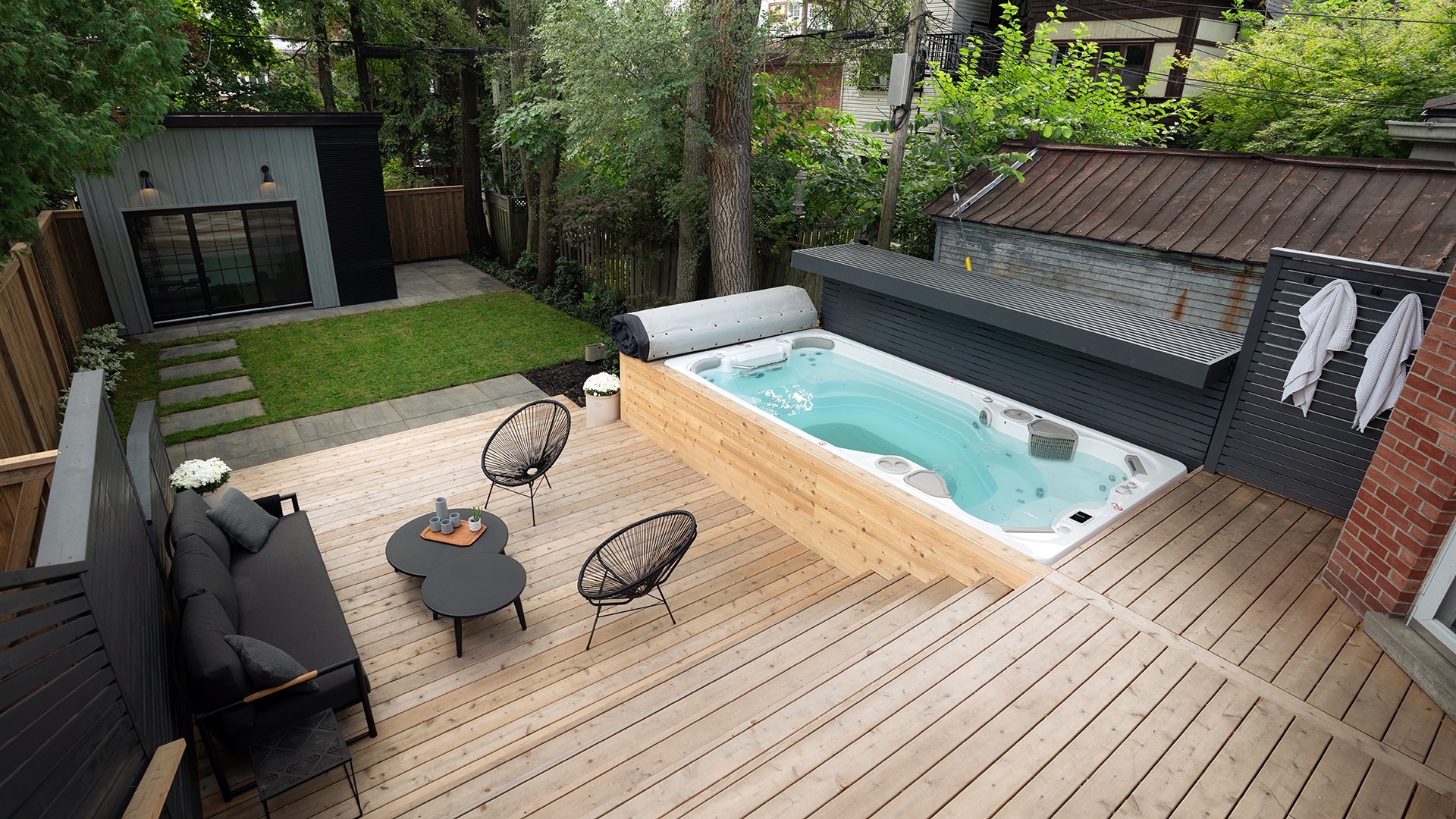 A modern backyard features a wooden deck with a hot tub, seating area, and lush greenery, creating a cozy outdoor relaxation space.