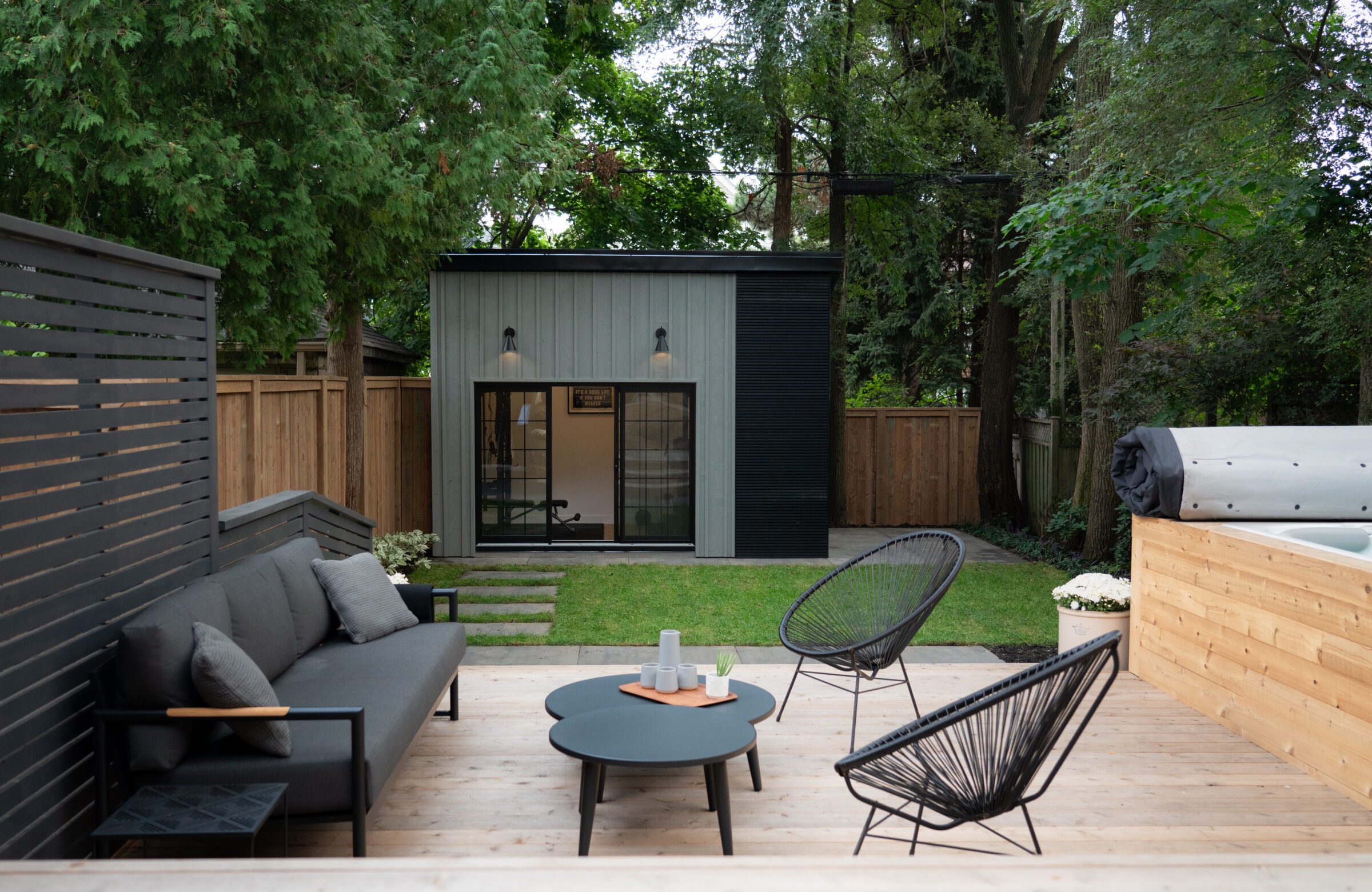 Modern backyard with a cozy patio, featuring a small building, outdoor seating, and surrounded by lush greenery and wooden fencing.